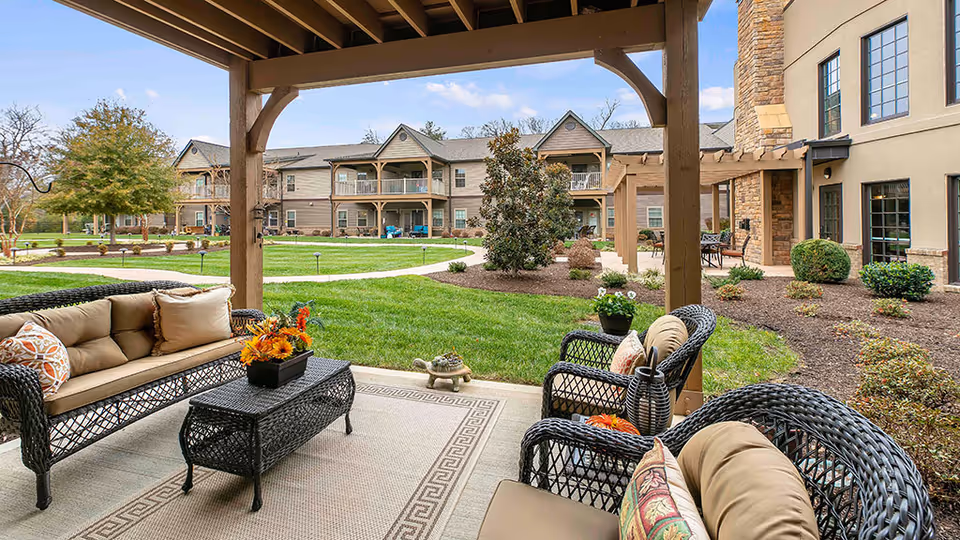 Outdoor seating area under a wooden pergola with wicker furniture including a sofa, chairs, and a coffee table with a flower arrangement. The seating area overlooks a well-maintained grassy courtyard with shrubs, trees, and a two-story building in the background.