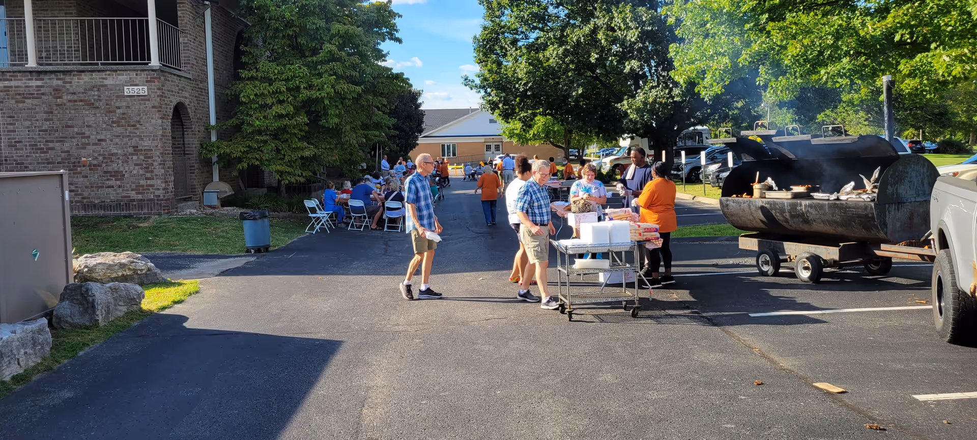 Outdoor gathering at Twinbrook Assisted Living Apartments with people socializing near a large barbecue grill in a parking lot. Several people are seated at tables in the background under trees, enjoying the event on a sunny day.