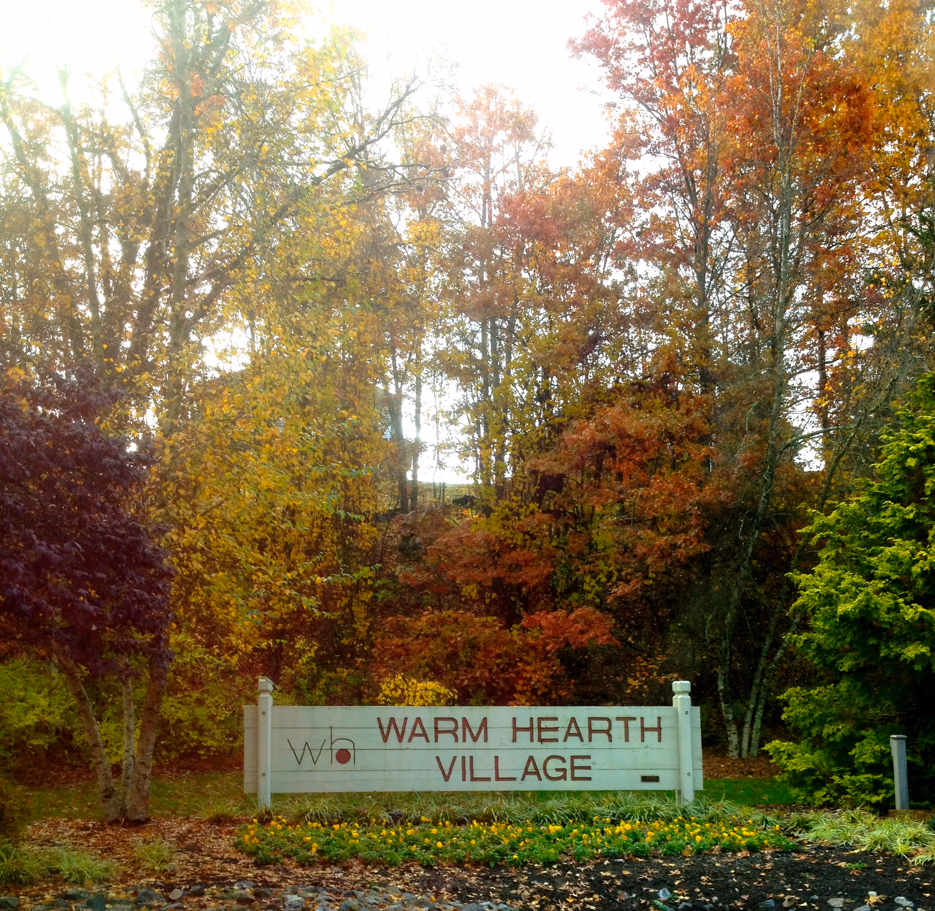 Outdoor scene showing a white sign with red letters that reads 'WARM HEARTH VILLAGE' surrounded by autumn trees with colorful leaves in shades of yellow, orange, and red.