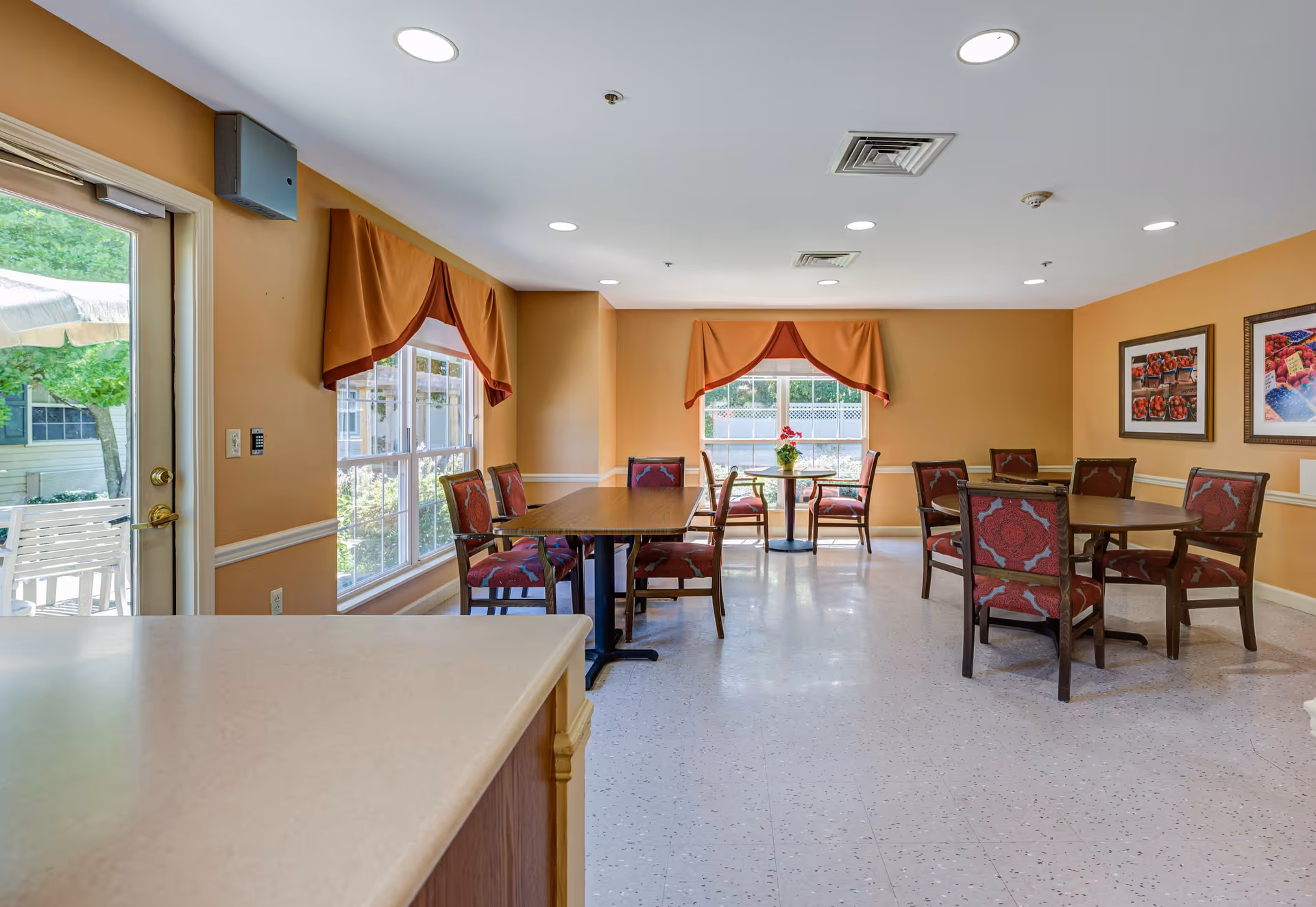 A bright dining room with several tables and chairs featuring red patterned upholstery. The walls are painted a warm beige with white trim, and there are large windows with orange valances letting in natural light. Two framed pictures hang on the right wall, and a door on the left leads outside to a patio area with a white bench and umbrella.