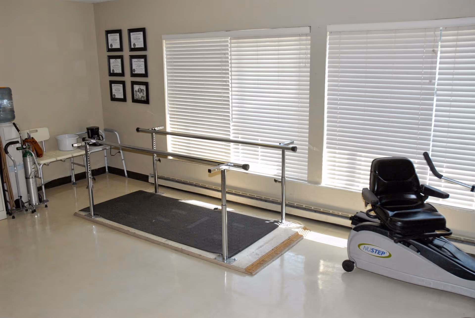 A rehabilitation room with parallel bars for walking exercises, a NuStep recumbent cross trainer, a water cooler, chairs, and framed certificates on the wall. The room has large windows with closed blinds letting in some natural light.