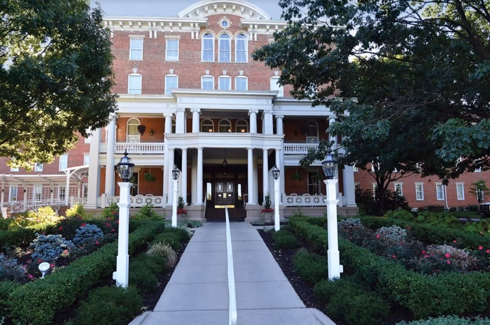 Front exterior view of a large, historic brick building with white columns and a covered entrance. The building is surrounded by well-maintained gardens with bushes, flowers, and trees. A concrete walkway with a handrail leads up to the entrance, flanked by white lamp posts.