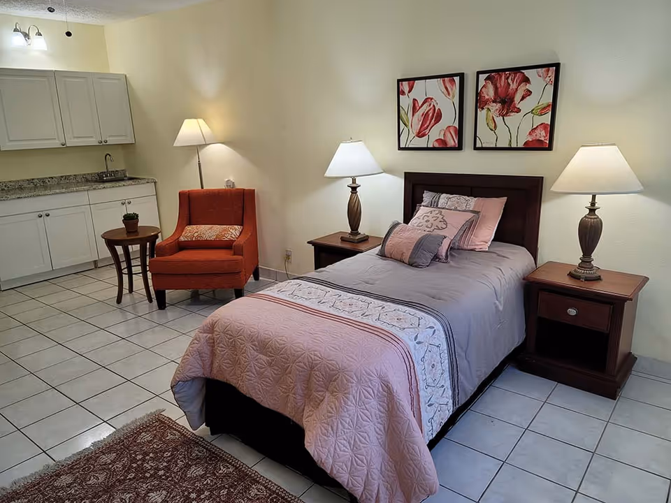 A cozy bedroom with a single bed covered in a pink and gray quilt, flanked by two wooden nightstands each with a lamp. Above the bed are two framed floral paintings. To the left, there is an orange armchair with a small round wooden table beside it. The room has white tiled floors and a kitchenette area with white cabinets and a granite countertop in the background.