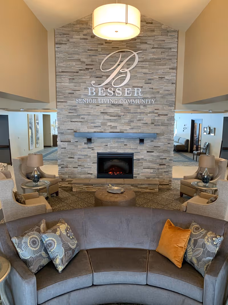 Lobby seating area with a curved sofa, armchairs, and a stone fireplace wall displaying the Besser Senior Living Community logo.