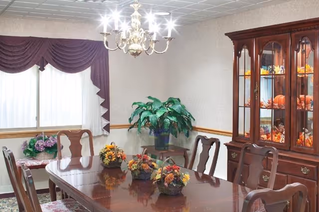 A traditional dining room with a polished wooden dining table surrounded by six matching chairs. The table is decorated with three floral centerpieces. A large wooden china cabinet filled with autumn-themed decorations stands against the wall. A green potted plant sits on a small side table in the corner. The room has a window with sheer white curtains and purple valances, and a brass chandelier hanging from the ceiling.