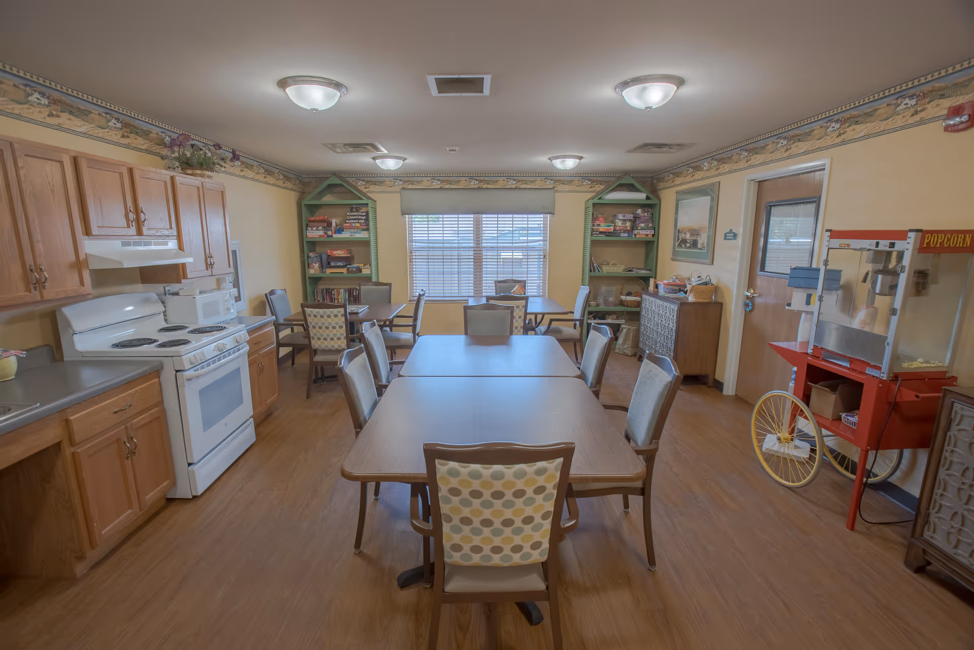 A communal kitchen and dining area with wooden cabinets, a white stove, microwave, and countertop on the left side. In the center, there are two rectangular tables pushed together with several chairs around them. At the back, there are two green shelves filled with board games and books. On the right side, there is a red popcorn machine on wheels and a door. The room has wood flooring and beige walls with a decorative border near the ceiling.