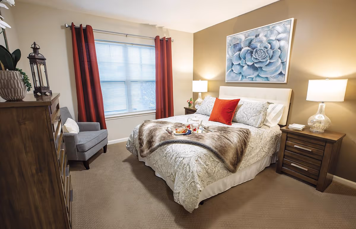 A cozy bedroom with a neatly made bed featuring patterned bedding, a fur throw, and a red accent pillow. There are two wooden nightstands with lamps on either side of the bed. A large window with red curtains lets in natural light. A gray armchair is positioned near a wooden dresser with decorative items on top. A large floral painting hangs above the bed.