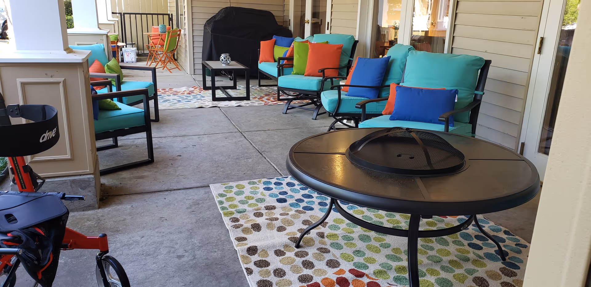 Outdoor patio area with turquoise cushioned chairs and colorful pillows arranged around a round fire pit table on a polka dot patterned rug. In the background, there is a covered grill, a small black coffee table, and additional seating with orange and green chairs. A red walker is partially visible on the left side.