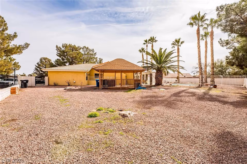 A single-story yellow building with a covered patio surrounded by a large gravel yard and palm trees.