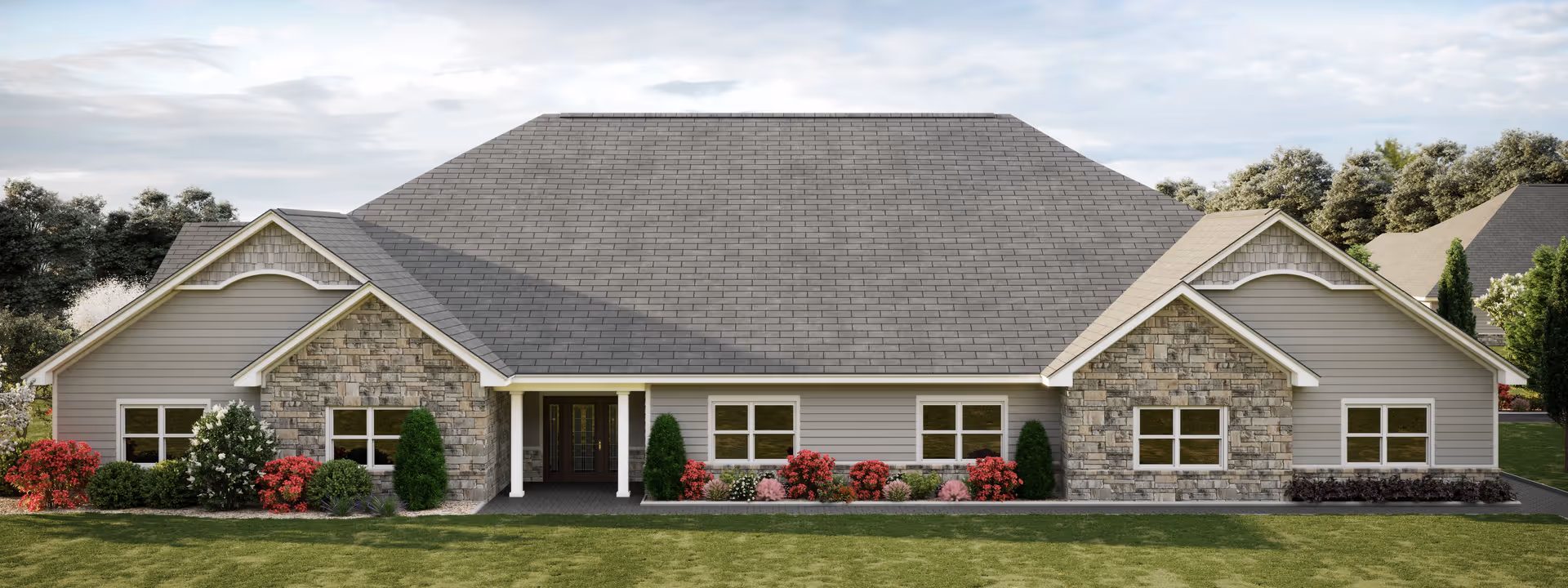 Front exterior view of a single-story residential building with a large gray shingled roof, stone and gray siding walls, white trim, and a central entrance with double doors. The building is surrounded by green grass, bushes, and colorful flowering plants.