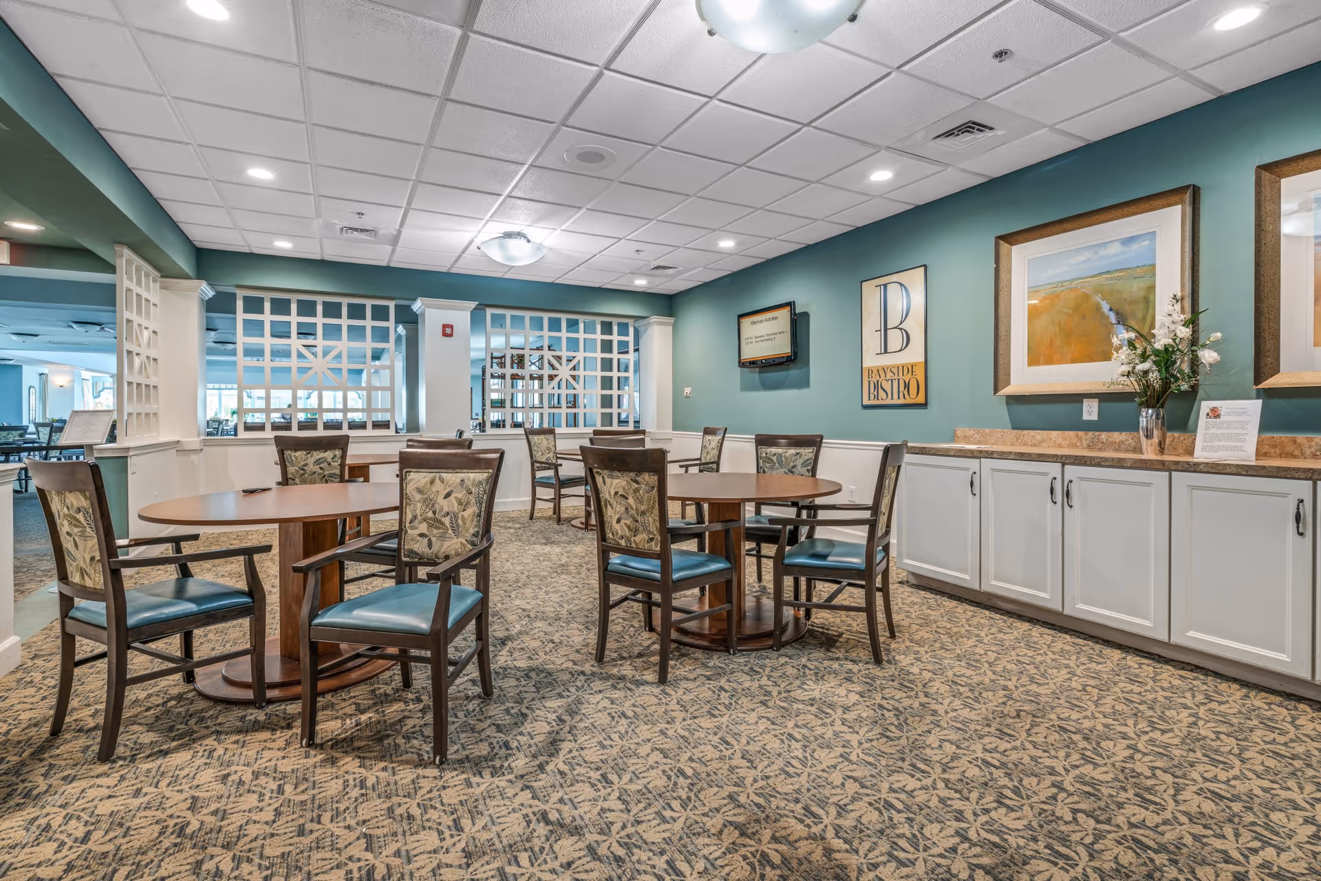 Interior view of a dining area with round wooden tables and chairs with patterned upholstery. The walls are painted teal with white wainscoting, decorated with framed artwork and a sign that reads 'Bayside Bistro'. There is a countertop with cabinets underneath on the right side and a flat-screen TV mounted on the wall. The ceiling has recessed lighting and a central light fixture.