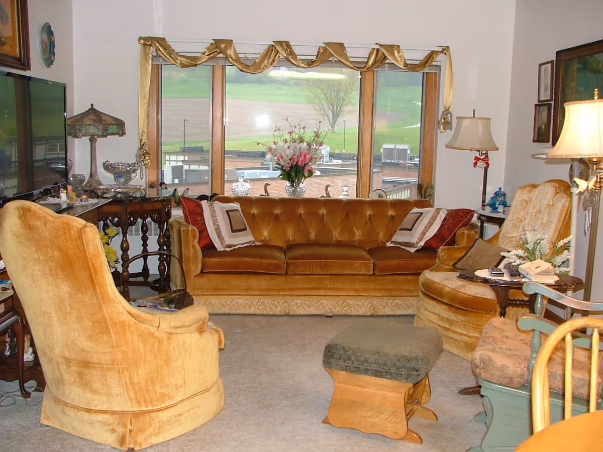 A cozy living room with vintage-style furniture including a mustard yellow sofa, matching armchairs, a green cushioned footstool, and a wooden side table with a lamp. There is a large window with a gold valance showing an outdoor view of a grassy area and some buildings. Various decorative items and framed pictures are on the walls and tables.