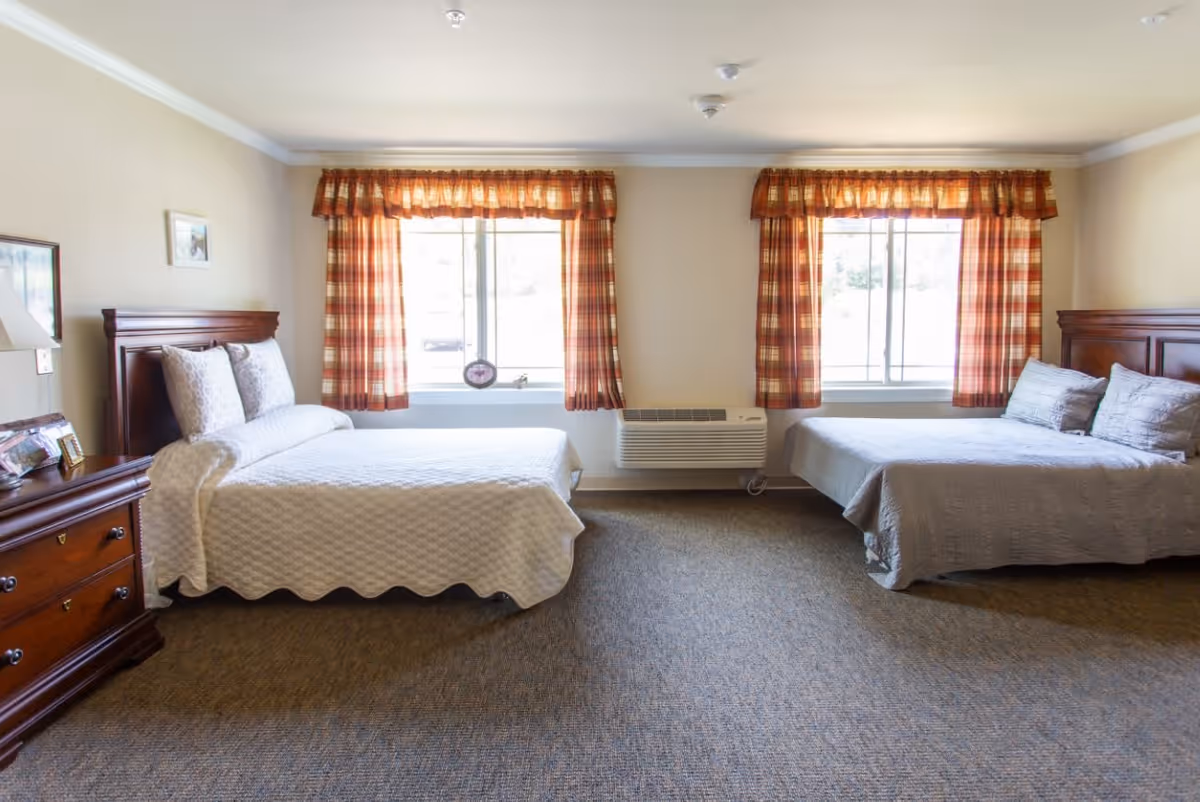 A bright bedroom with two beds, each with wooden headboards and light-colored bedding. There are two windows with plaid curtains letting in natural light. A wooden dresser with a lamp and framed photos is on the left side of the room. The floor is carpeted and there is an air conditioning unit mounted below the windows.
