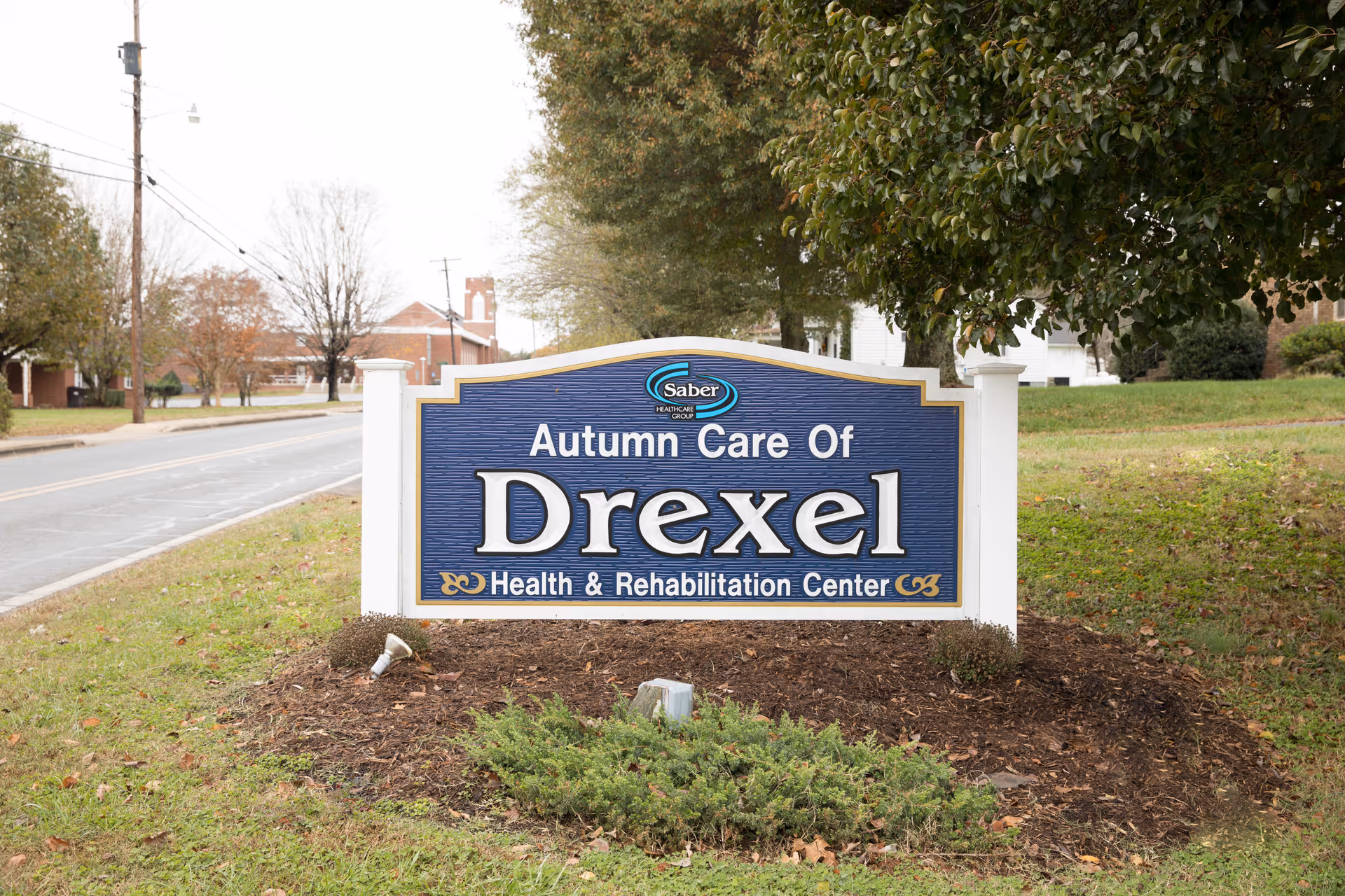 A blue and white sign for Autumn Care of Drexel Health & Rehabilitation Center located on a grassy area beside a road with trees and buildings in the background.