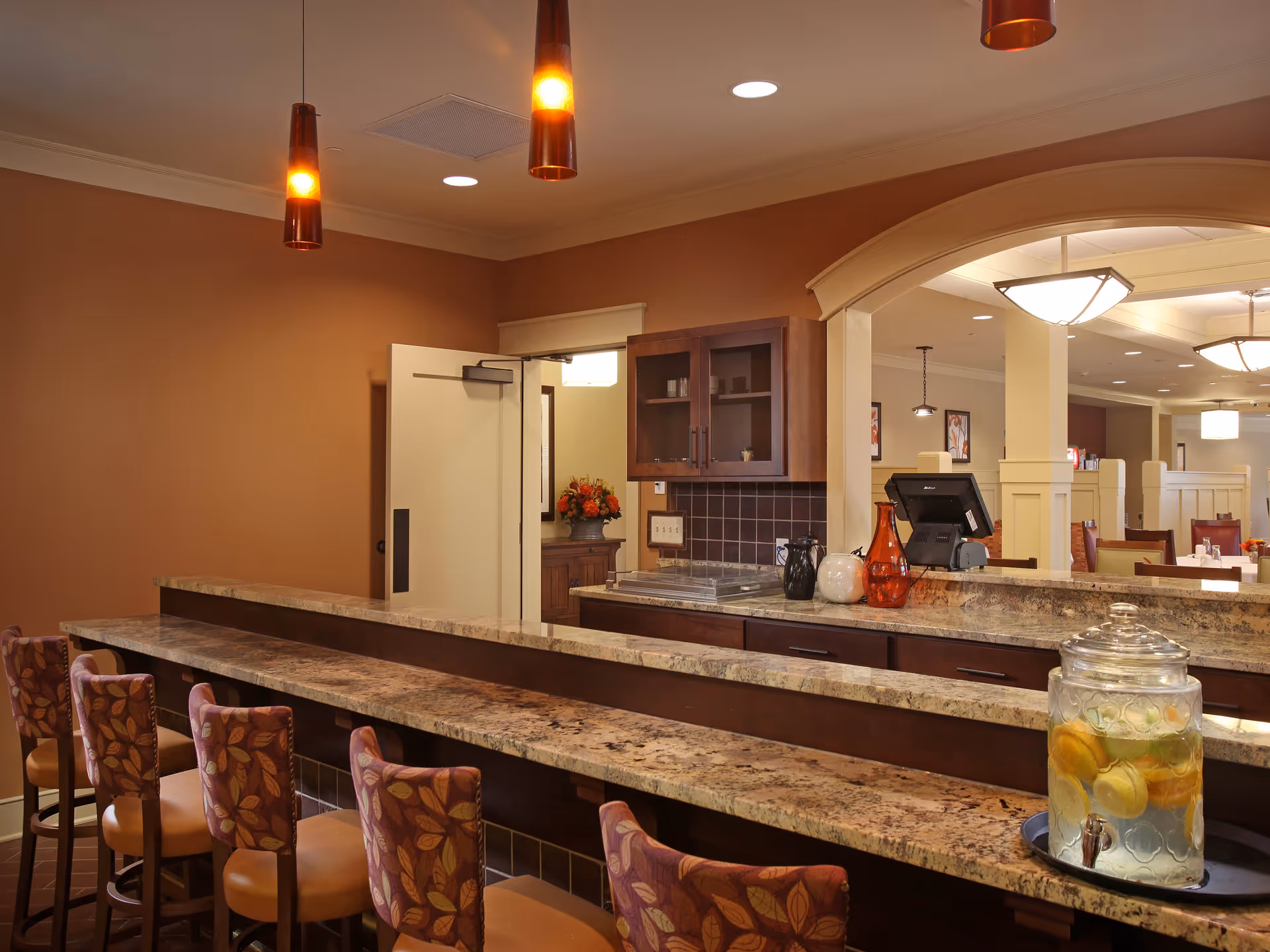 Interior view of a dining area with a long granite countertop bar and several high chairs with patterned upholstery. Behind the counter, there is a cabinet with glass doors, decorative vases, and a glass dispenser filled with lemon water. Warm lighting fixtures hang from the ceiling, and an open doorway leads to another room with additional seating and soft lighting.