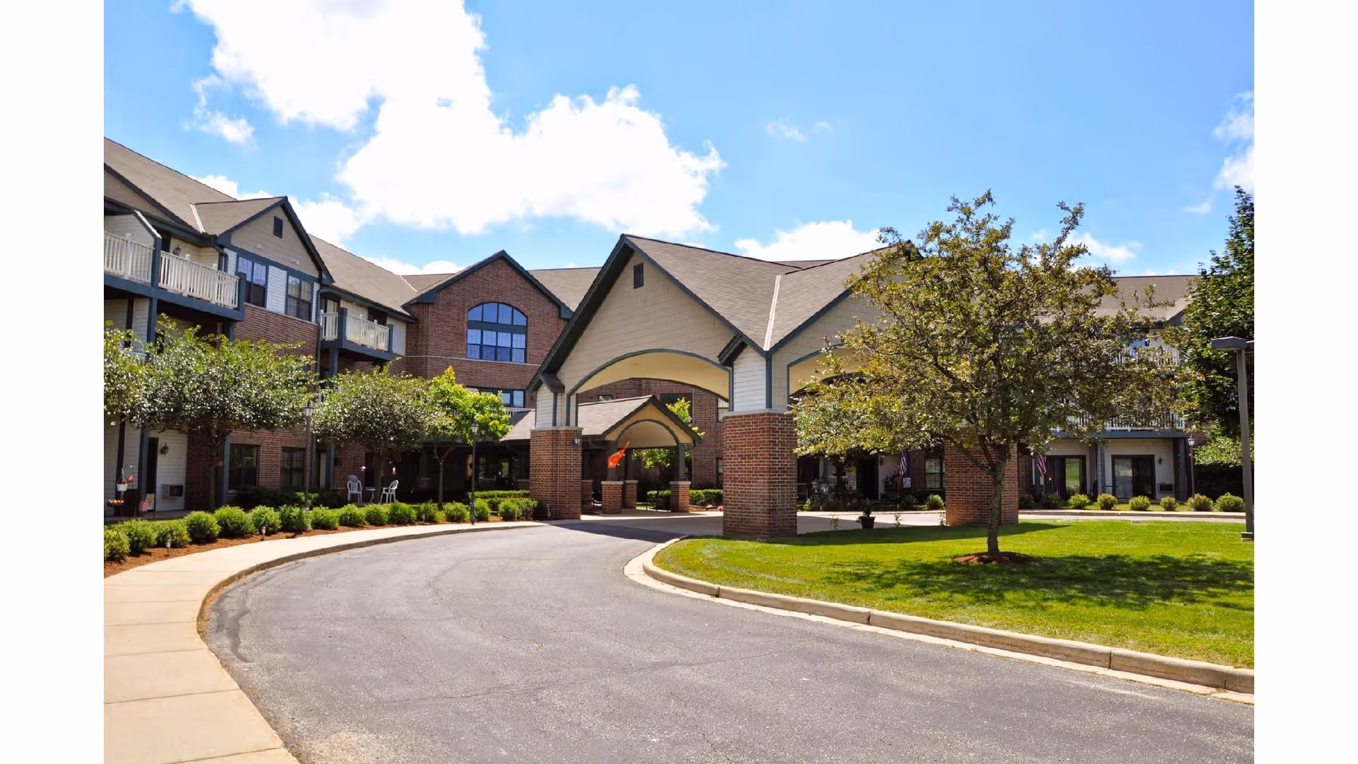 Exterior view of a senior living facility with a curved driveway leading to a covered entrance. The building has brick and siding walls with multiple windows and balconies. There are trees, shrubs, and a well-maintained lawn surrounding the entrance area under a partly cloudy sky.