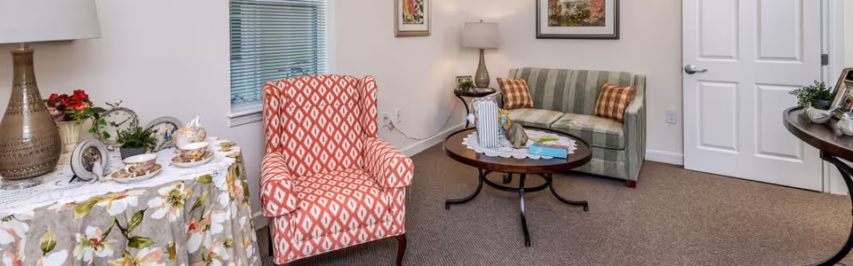 Small sitting area with a patterned armchair and striped loveseat around a round coffee table, with side tables holding a lamp and a tea set.