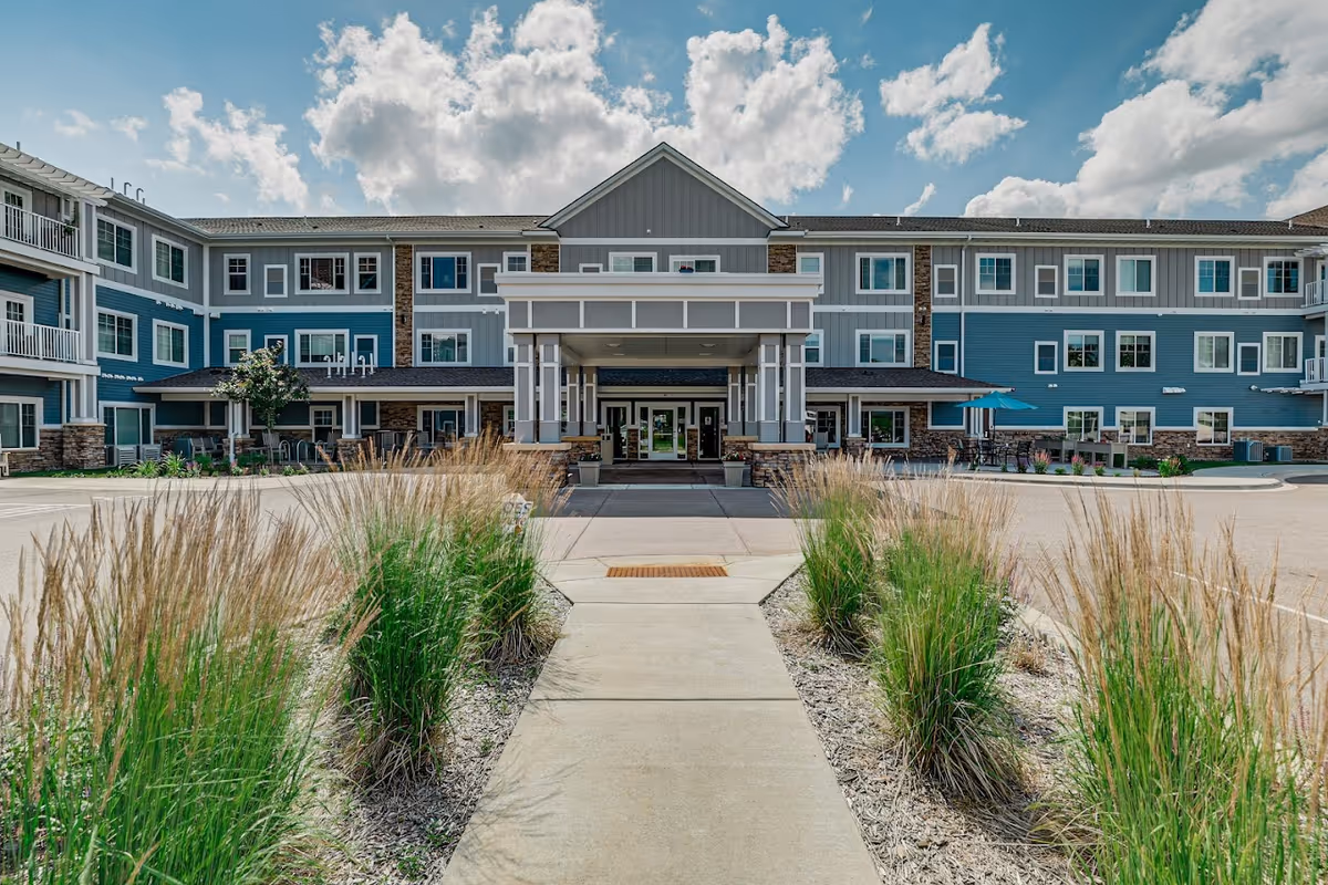 Front exterior view of a three-story senior living facility building with blue and gray siding, white trim, and stone accents. A concrete walkway leads to a covered entrance with pillars. Ornamental grasses line the walkway, and there are outdoor seating areas with umbrellas on the right side. The sky is partly cloudy with bright sunlight.