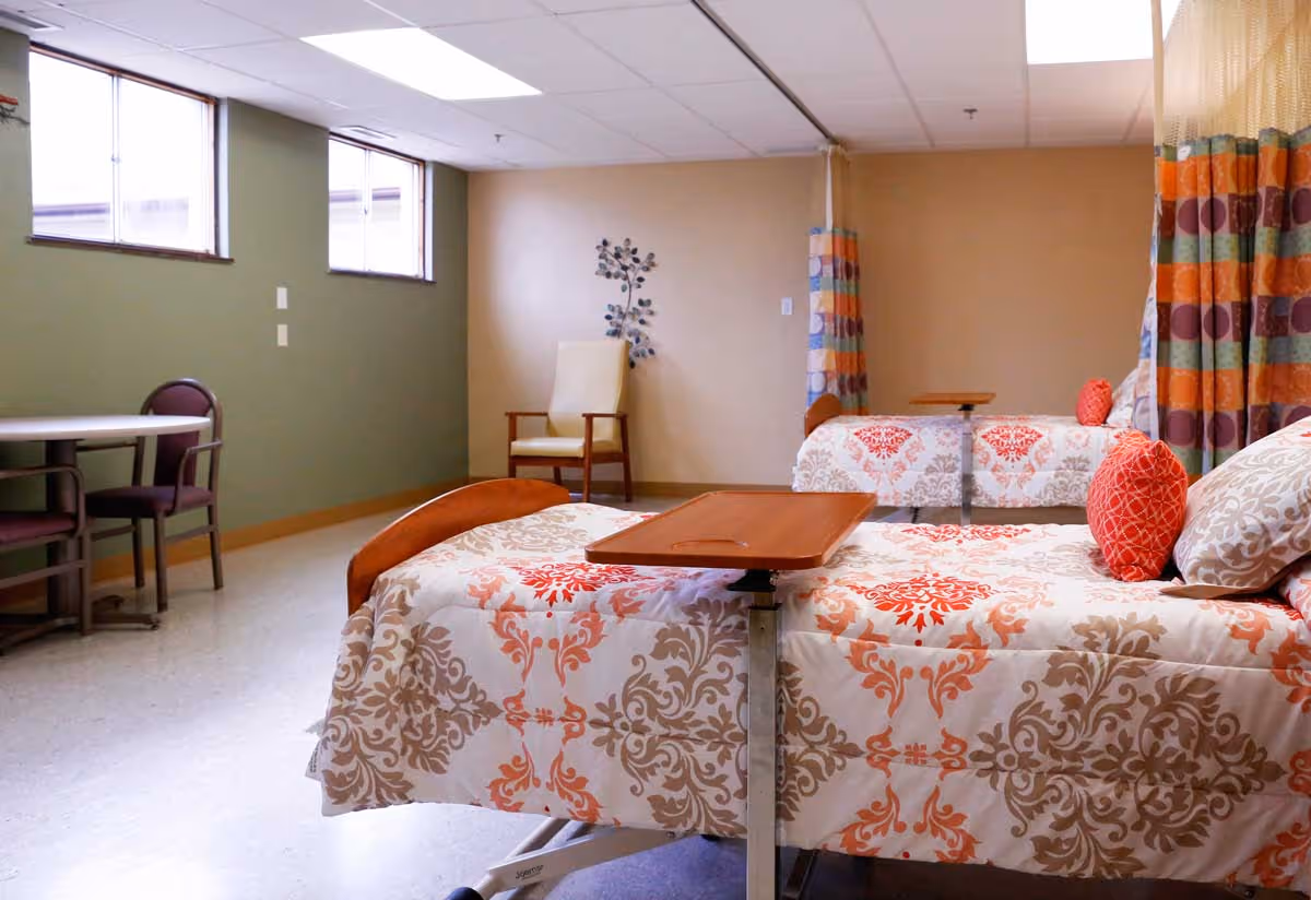 A room with two hospital-style beds covered in patterned bedspreads with red and beige designs, each with a red decorative pillow. There is a wooden overbed table on the nearest bed. The room has a beige wall with a decorative metal wall art piece, a beige armchair, a small round table with two chairs near windows, and colorful curtains with orange, green, and purple stripes.