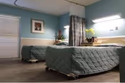 Two twin nursing beds in a softly lit patient room with a curtain divider, window blinds, and a bedside table.