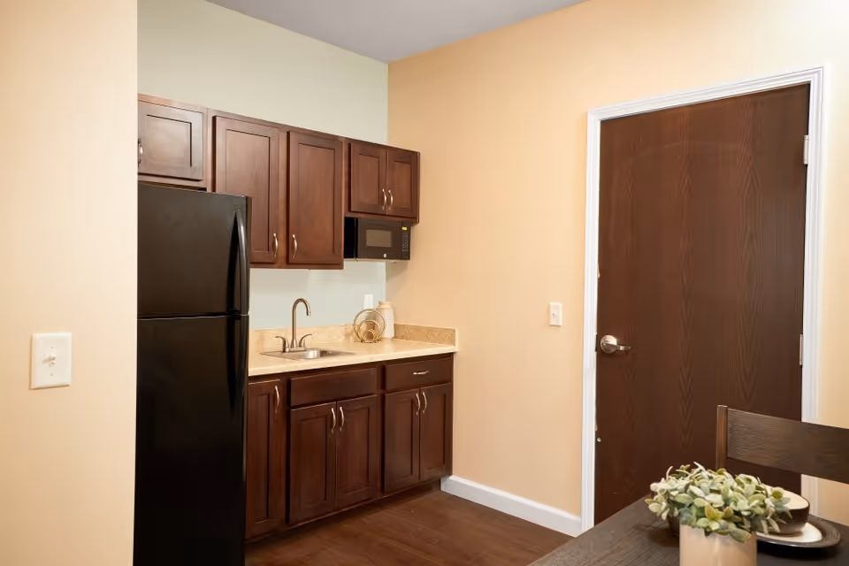 A small kitchen area with dark wooden cabinets, a black refrigerator, a microwave, a sink with a faucet, and a beige countertop. There is a dark wooden door on the right side and part of a dining table with a small potted plant and dishware visible in the foreground.