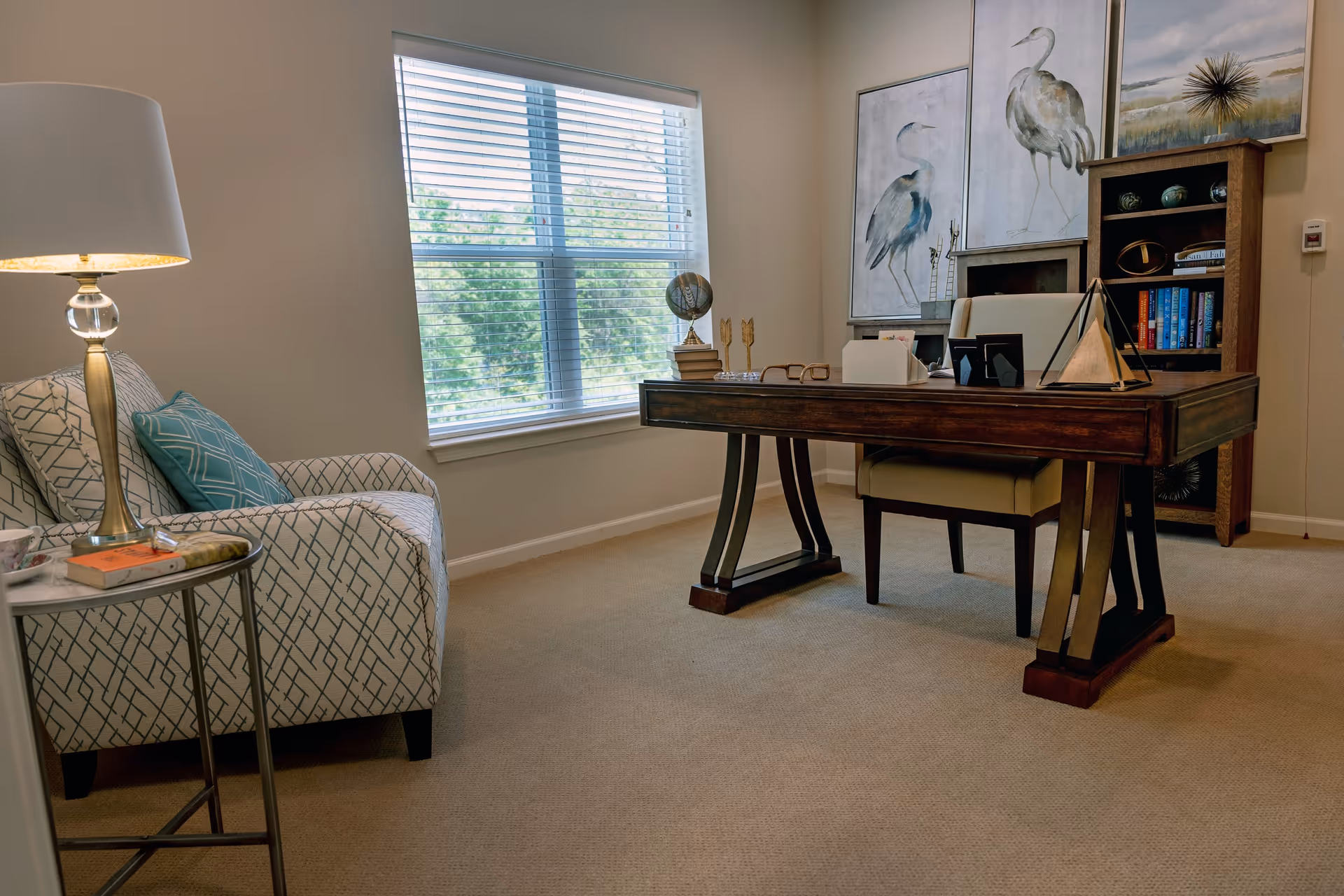 A cozy office space with a wooden desk and beige chair, a patterned armchair with blue and white cushions, a side table with a lamp and books, a window with blinds letting in natural light, and a bookshelf with books and decorative items. Two large paintings of birds hang on the wall behind the desk.