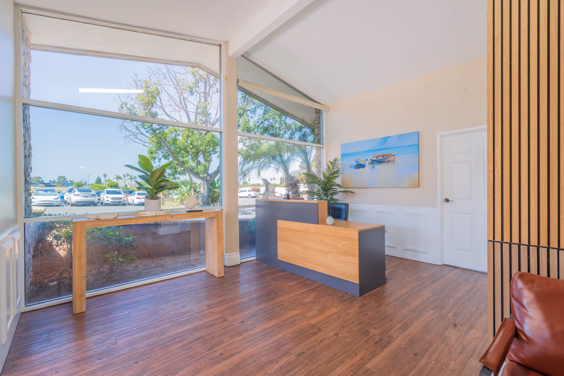 Bright reception area with large floor-to-ceiling windows showing a parking lot and trees outside. Inside, there is a modern wooden reception desk with a plant on it, a wooden console table with a plant and decorative items, a painting of boats on the wall, and wood flooring.
