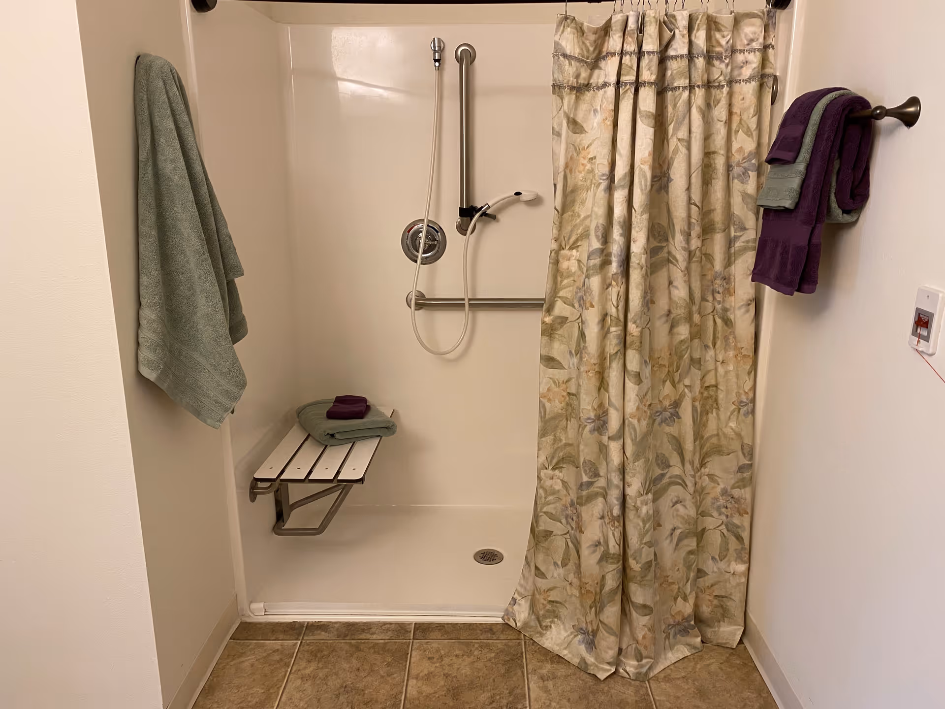 A walk-in shower with a floral patterned shower curtain, a fold-down white shower bench with folded green and purple towels on it, a handheld showerhead attached to a grab bar, and additional green and purple towels hanging on towel racks on either side of the shower.