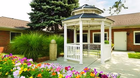 A white wooden gazebo with a bench inside situated on a concrete patio area surrounded by colorful flowers and green shrubs, with a brick building and large evergreen tree in the background under a partly cloudy sky.