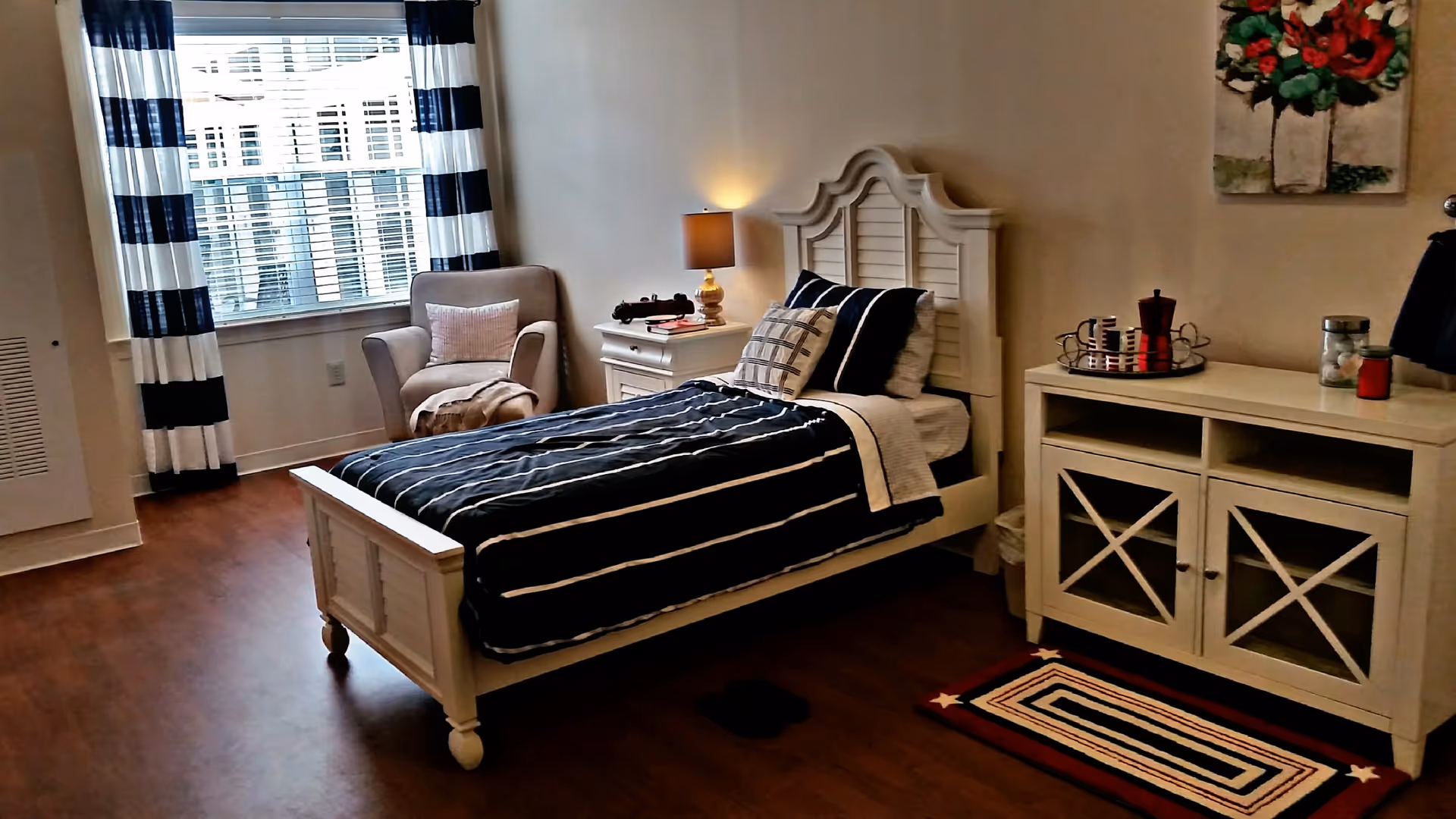 A cozy bedroom with a single bed featuring a white wooden frame and navy blue striped bedding. Next to the bed is a white nightstand with a lamp and decorative items. A beige armchair with a pillow and blanket is placed near a window with white blinds and navy and white striped curtains. Across from the bed is a white cabinet with glass-paneled doors, a tray with cups, and a red coffee maker on top. A colorful rug with stars is on the wooden floor, and a floral painting hangs on the wall.