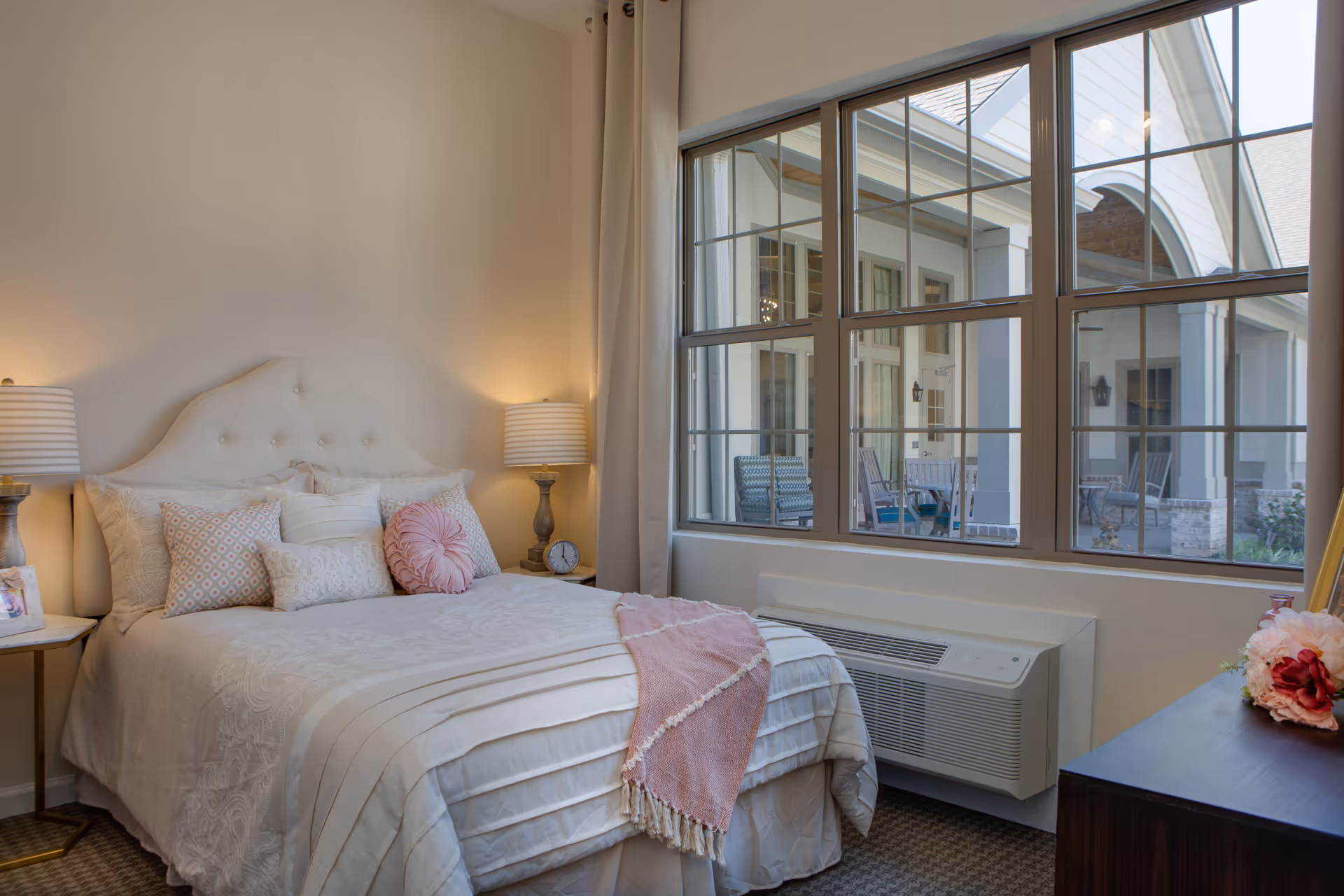 A cozy bedroom with a neatly made bed featuring multiple decorative pillows and a pink throw blanket. Two bedside tables each hold a lamp with a striped shade, and there is a clock on the right table. Large windows on the right side of the room offer a view of an outdoor patio area with chairs and tables. The room has soft, neutral-colored walls and carpeted flooring.