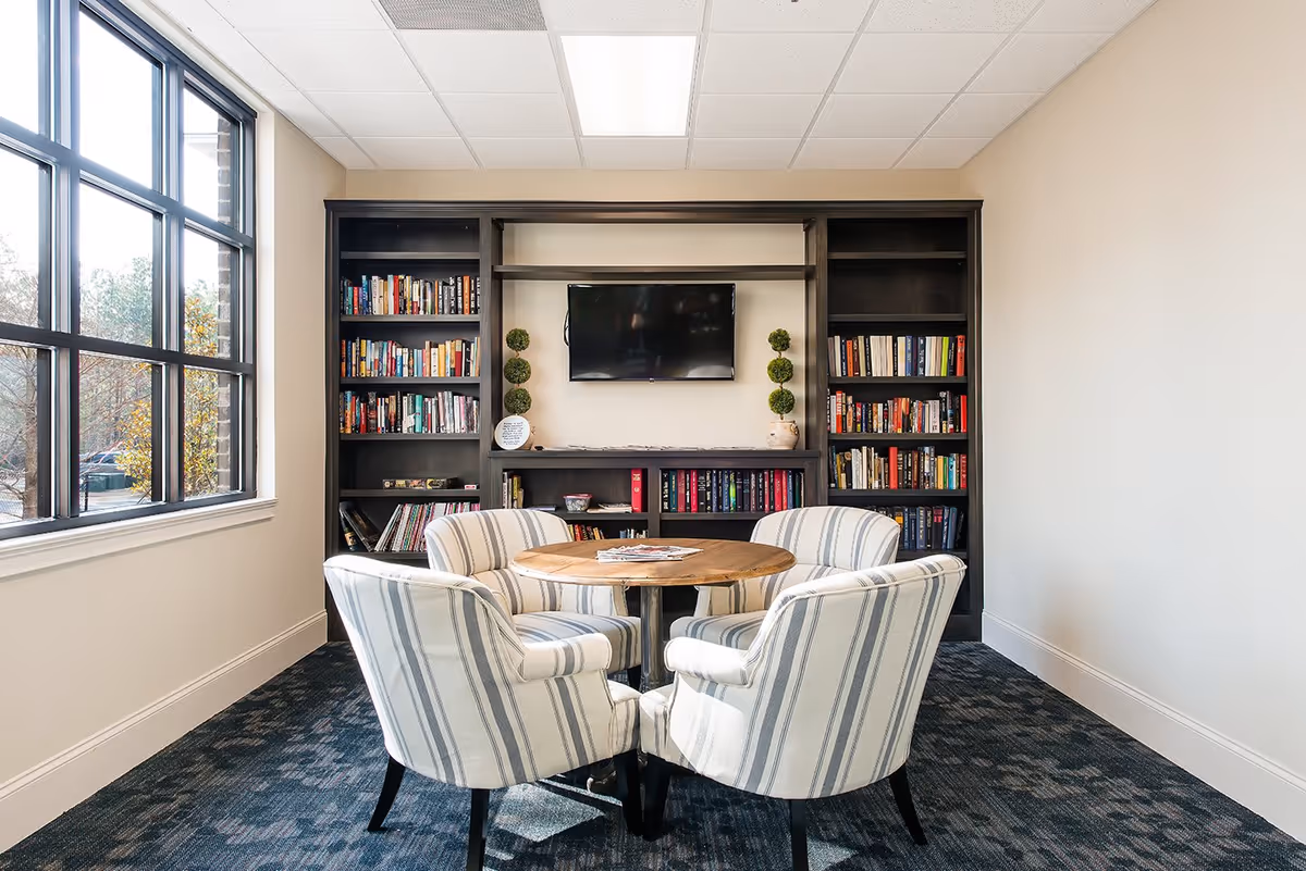 A cozy reading or meeting area with four striped upholstered armchairs arranged around a round wooden table. Behind the table is a large dark wood bookshelf filled with books and decorative items, and a flat-screen TV mounted on the wall. Large windows on the left side let in natural light, illuminating the room.