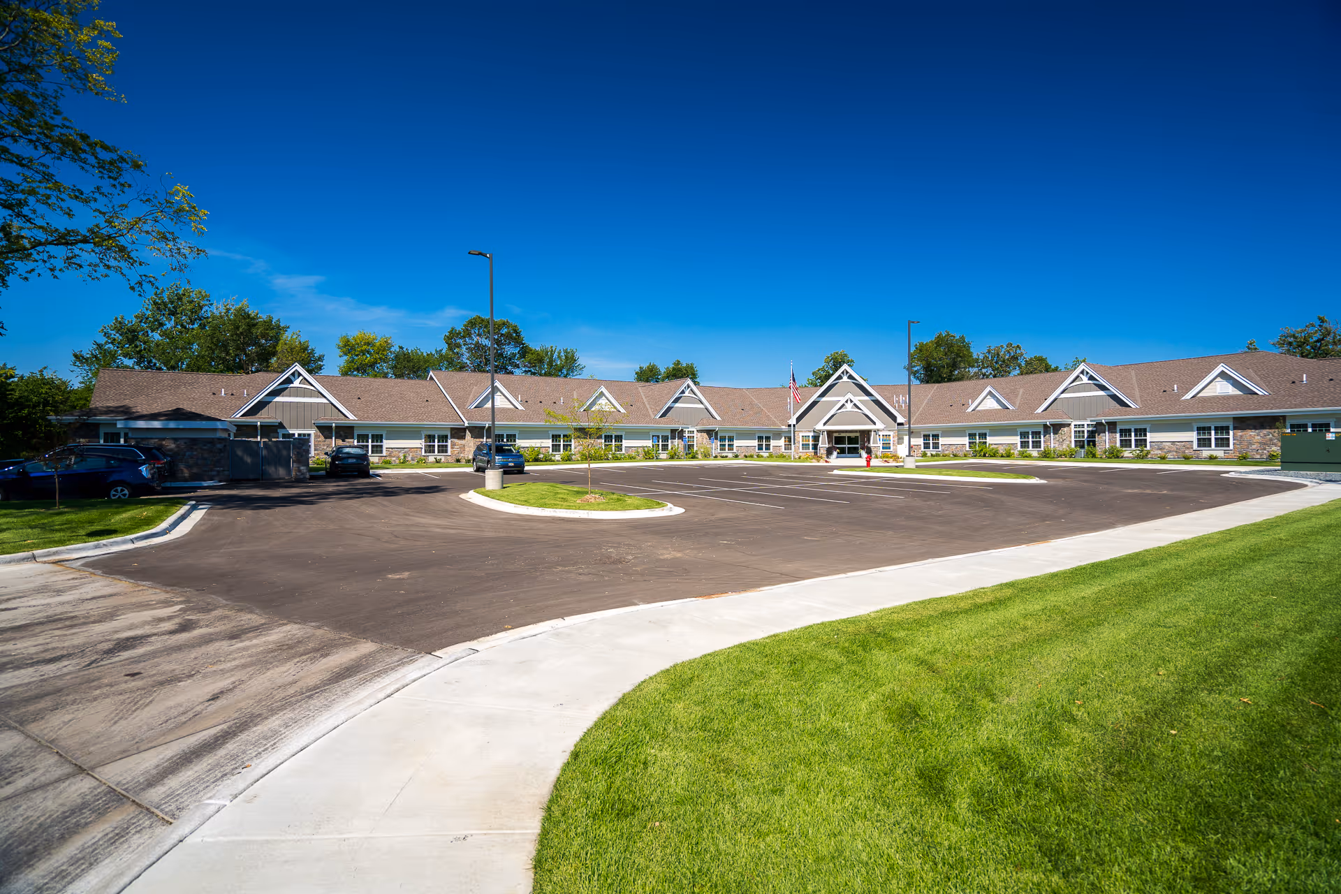 Wide exterior view of NorBella Senior Living - Champlin building under a clear blue sky, showing a large parking lot in front, green grass on the sides, and several cars parked near the building.