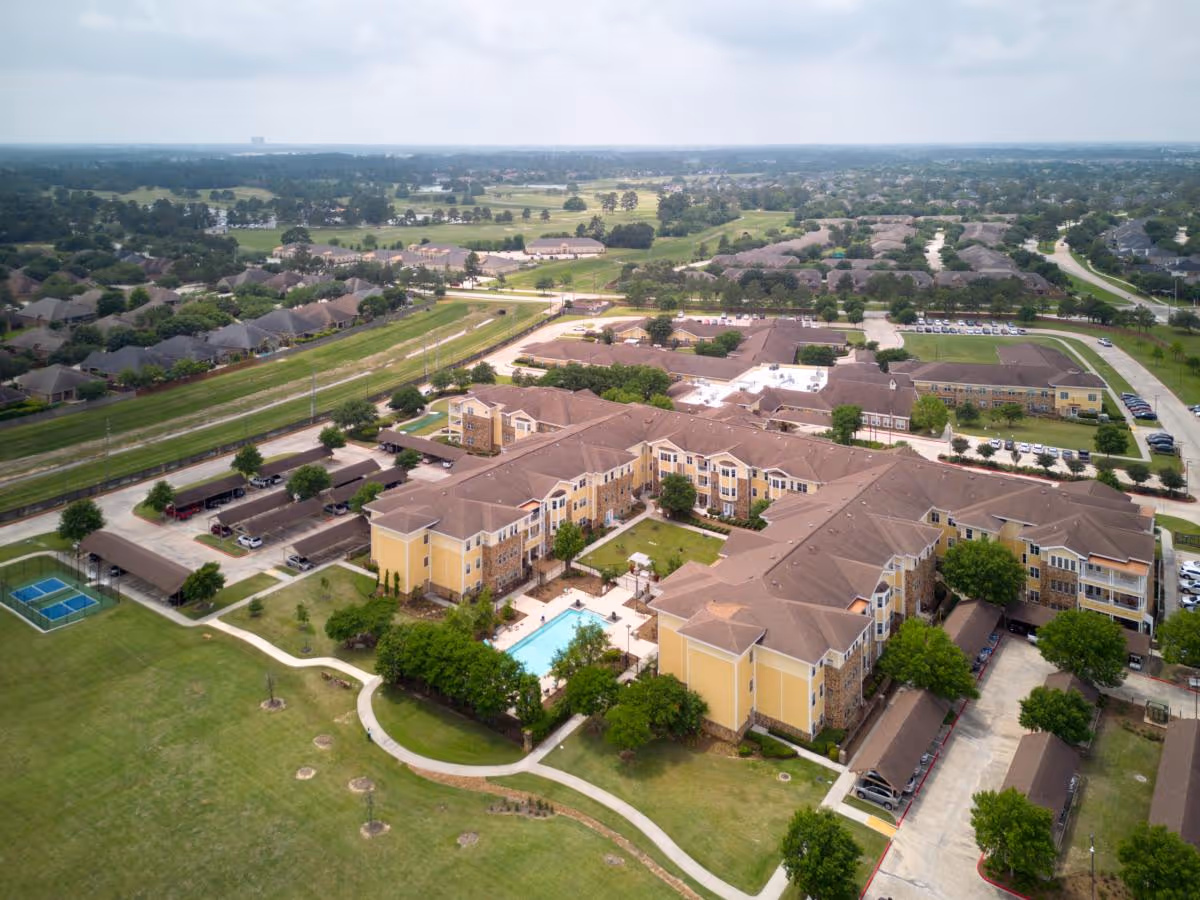 Aerial view of The Village at Gleannloch Farms senior living facility showing multiple connected yellow buildings with brown roofs surrounding a central courtyard with a swimming pool. The facility is surrounded by green lawns, trees, parking areas, and nearby residential neighborhoods under a cloudy sky.