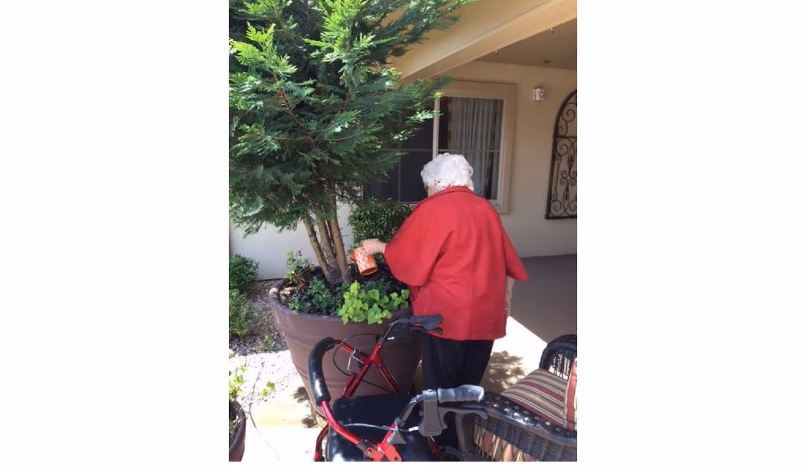 A person in a red jacket tending plants in a large planter on a covered patio with a walker and outdoor chairs nearby.