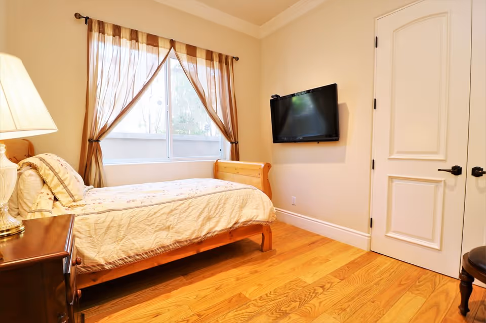 A cozy bedroom with a single wooden bed covered with a light-colored quilt and pillows. There is a wooden nightstand with a lamp on the left side of the bed. A window with sheer brown curtains lets in natural light. A flat-screen TV is mounted on the wall opposite the bed, and there is a white door with black handles on the right side. The floor is made of light wood.