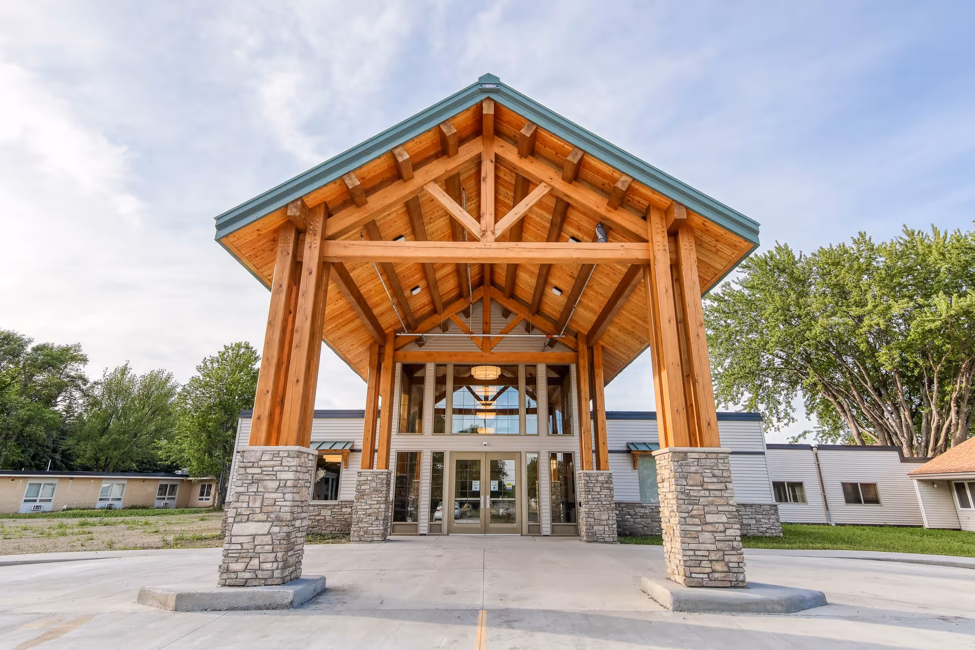 Front exterior view of Maplewood Manor Senior Living facility featuring a large wooden entrance structure with stone pillars and glass doors under a partly cloudy sky.