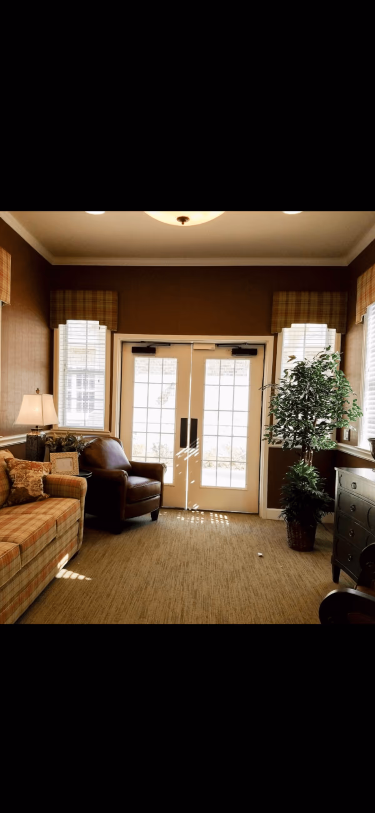 A cozy living room area with a plaid sofa on the left, a brown leather armchair next to it, and a large potted plant on the right near a window. Double glass doors with white frames are centered in the background, flanked by two windows with plaid valances. A lamp on a side table is next to the sofa, and a dark wooden dresser is on the right side of the image.