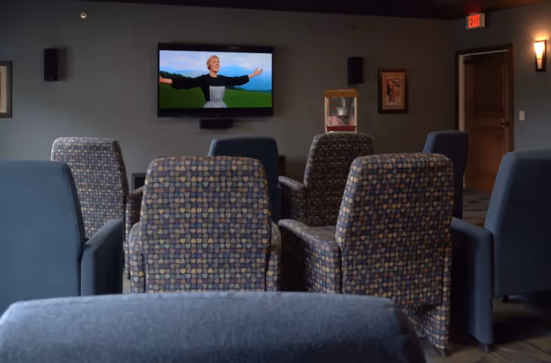 A cozy senior living common area with several patterned and solid blue armchairs arranged facing a wall-mounted television showing a scene from The Sound of Music. The room has dim lighting, a popcorn machine in the corner, framed artwork on the walls, and an exit door with a red exit sign above it.