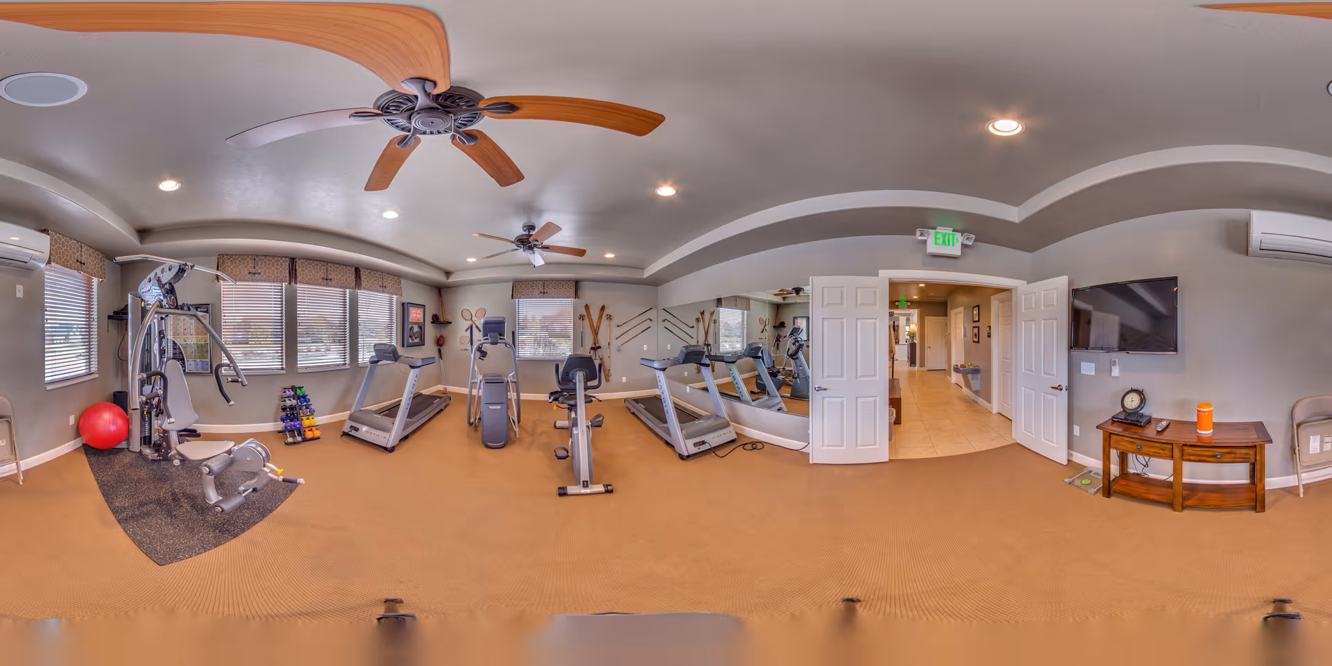 A fitness room in Bridlewood Villas featuring exercise equipment including treadmills, a stationary bike, a multi-gym machine, and free weights. The room has beige carpet, gray walls, ceiling fans, and large windows letting in natural light. A TV is mounted on the wall above a small wooden table with a clock and water bottle. Double doors open to a hallway.