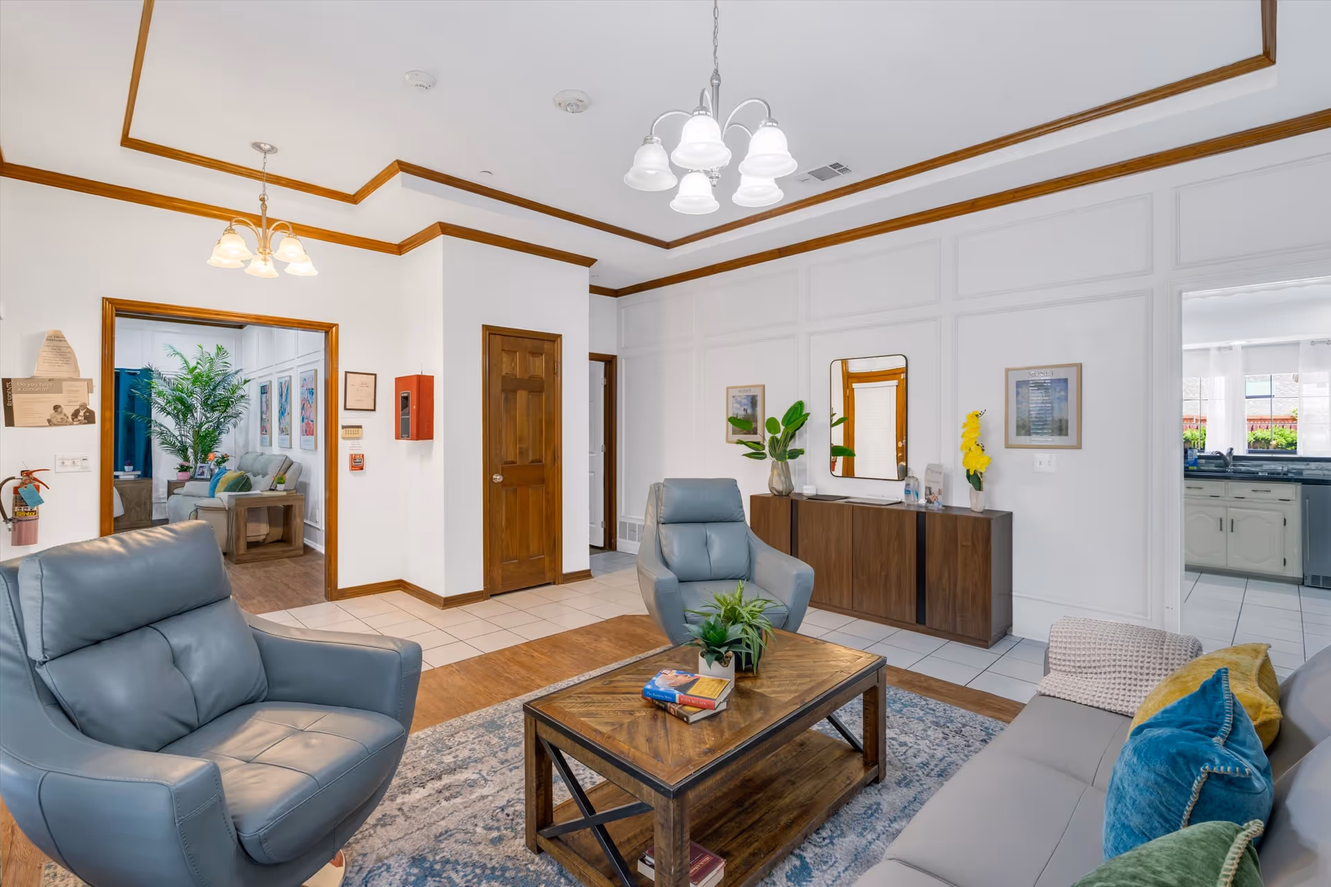 A bright and cozy living room area with two gray leather armchairs and a gray sofa adorned with colorful pillows. A wooden coffee table with books and a small plant sits on a patterned rug. The room features white walls with wooden trim, a wooden sideboard with decorative plants and a mirror above it. Two doorways lead to adjacent rooms, one showing a glimpse of a kitchen and the other a seating area with a large plant and artwork on the walls. Ceiling lights hang from the white ceiling with crown molding.