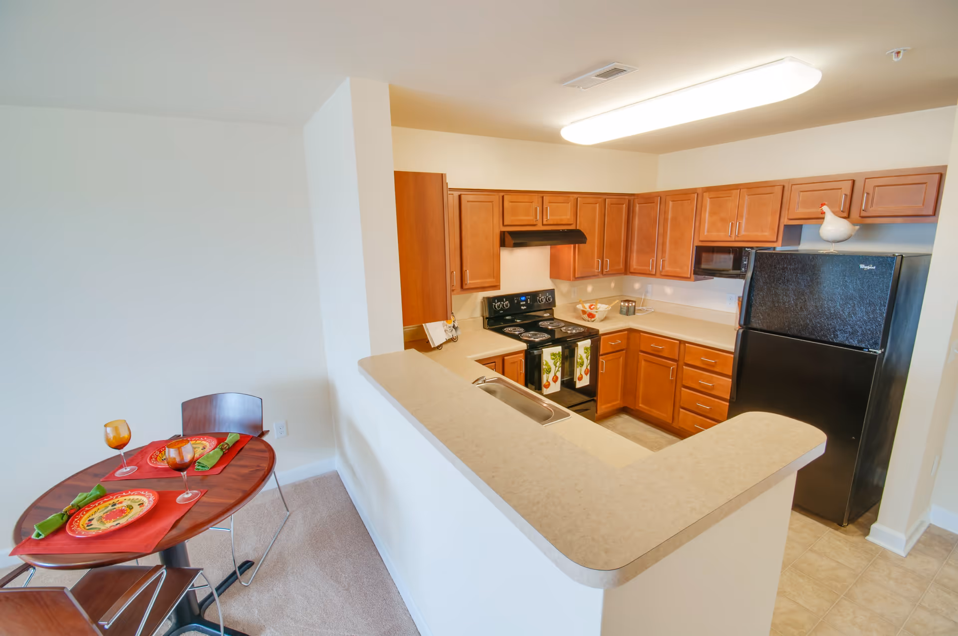 A compact kitchen with wooden cabinets, a black refrigerator, an electric stove with two vegetable-themed towels hanging on the oven door, a microwave, and a countertop with a built-in sink. Adjacent to the kitchen is a small dining area with a round wooden table set with two place settings, each with a colorful plate, green napkin, and amber-colored glass.