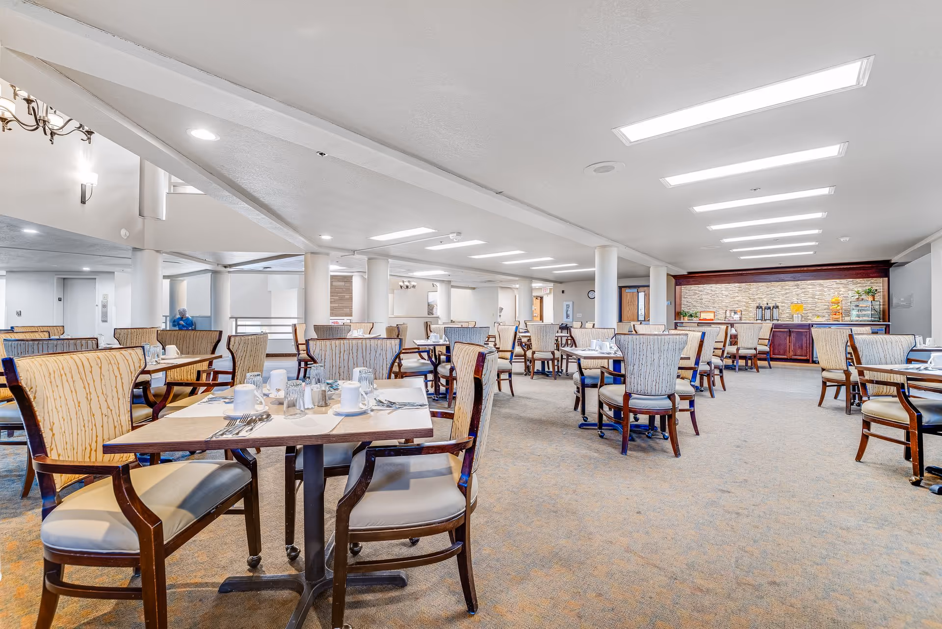 A spacious dining room in a senior living facility with multiple tables and chairs arranged neatly. Each table is set with cups, saucers, and silverware. The room has bright overhead lighting, carpeted floors, and a buffet area with beverages and snacks at the far end.