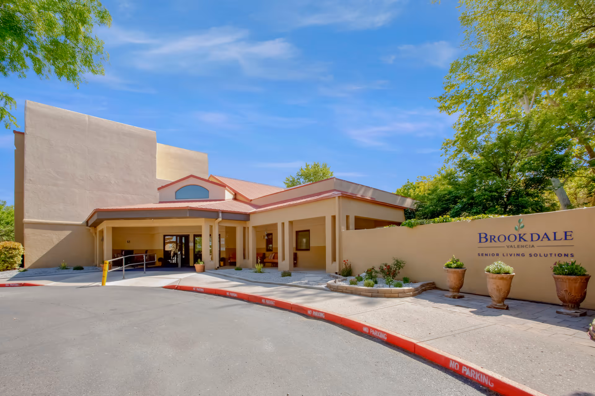 Exterior view of the Brookdale Valencia senior living facility entrance with a covered drop-off area, beige building walls, potted plants, and a clear blue sky.