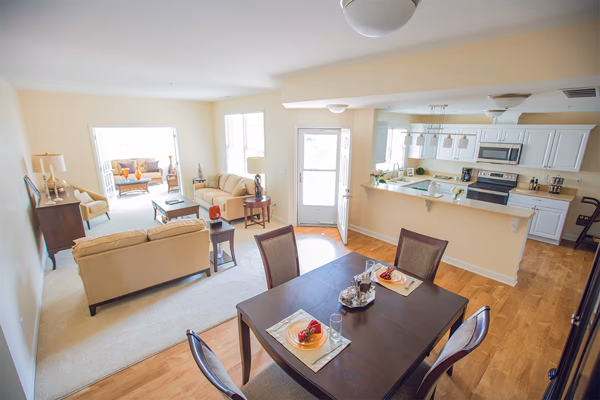 Bright and spacious open-concept living area in a senior living facility with a dining table set for two in the foreground, a kitchen with white cabinets and modern appliances to the right, and a cozy living room with beige sofas and a coffee table in the background. Large windows and a glass door allow natural light to fill the space.