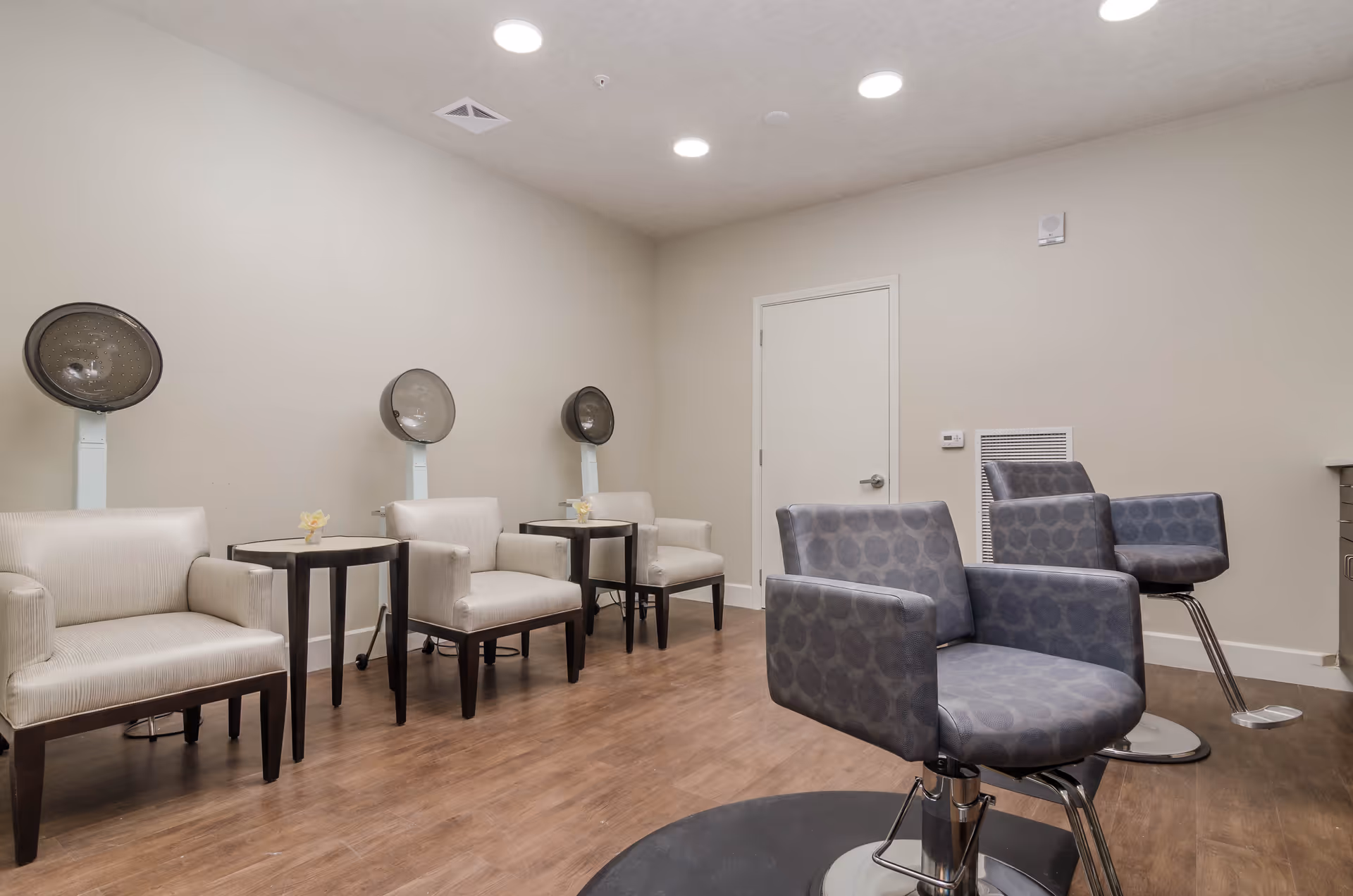 Interior of a salon area in a senior living facility with three beige chairs each paired with a hair dryer, two gray salon chairs on hydraulic bases, wooden flooring, and a closed white door in the background.