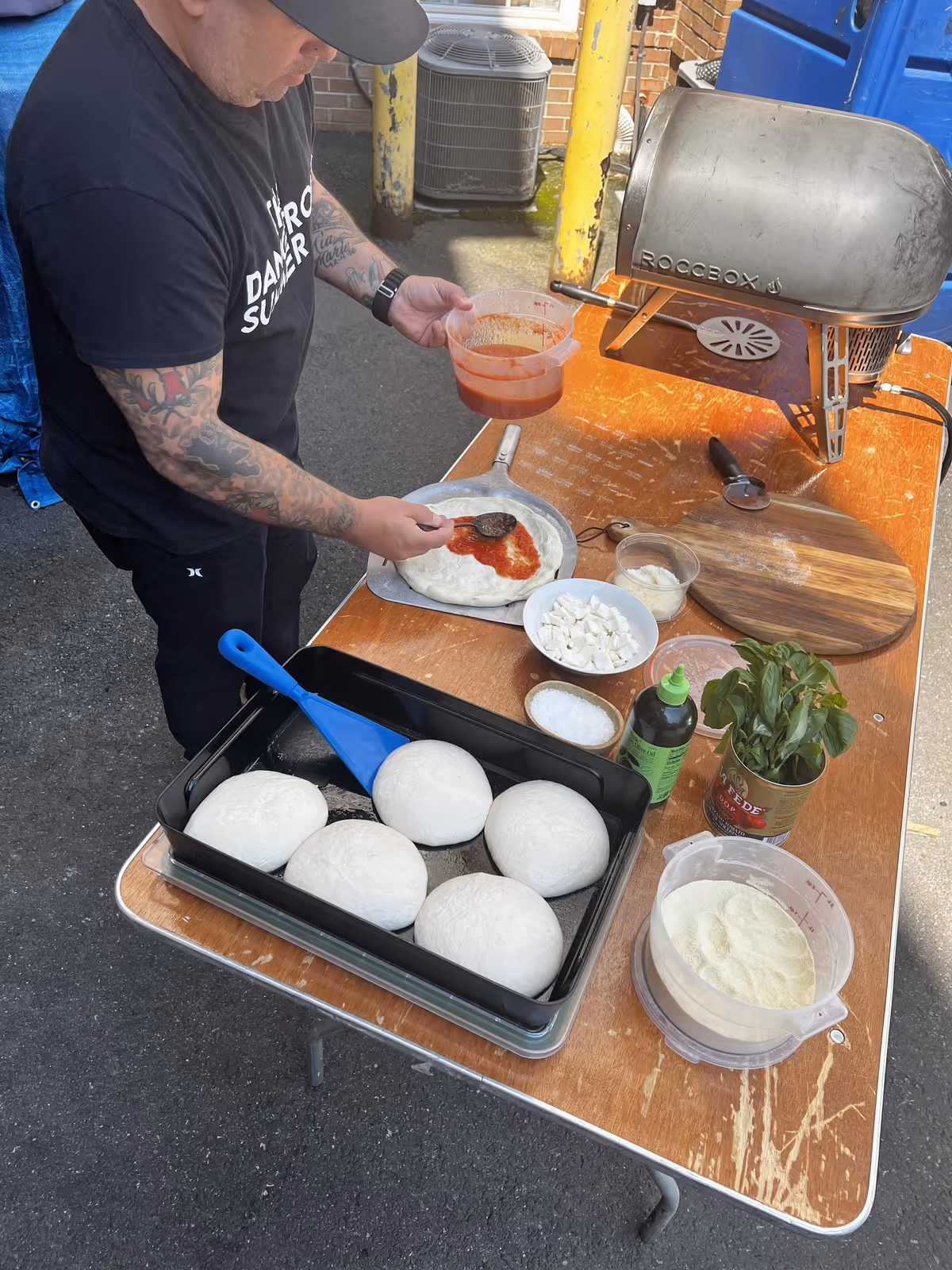A person with tattooed arms and wearing a black t-shirt is spreading tomato sauce on a pizza dough placed on a pizza peel. On the table, there are several balls of pizza dough in a black tray, a bowl of cubed cheese, a bowl of salt, a container of green sauce, a bunch of fresh basil in a can, a wooden cutting board, and a pizza oven labeled 'Roccbox'. The setting appears to be outdoors on a paved surface.
