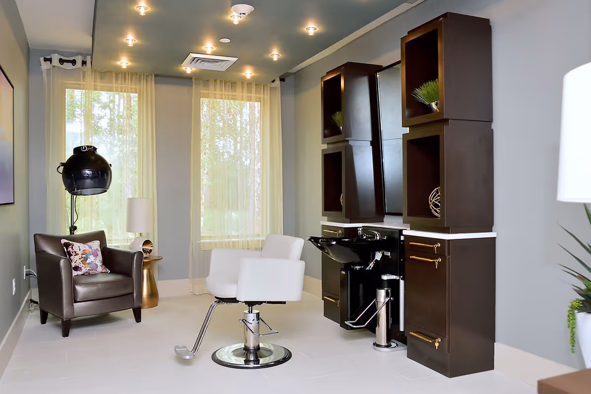 Interior of a senior living facility hair salon with a white salon chair in front of a black hair washing station and dark wood cabinets. There is a brown leather armchair with a floral pillow and a gold side table with a lamp next to it. Two large windows with sheer curtains allow natural light into the room.