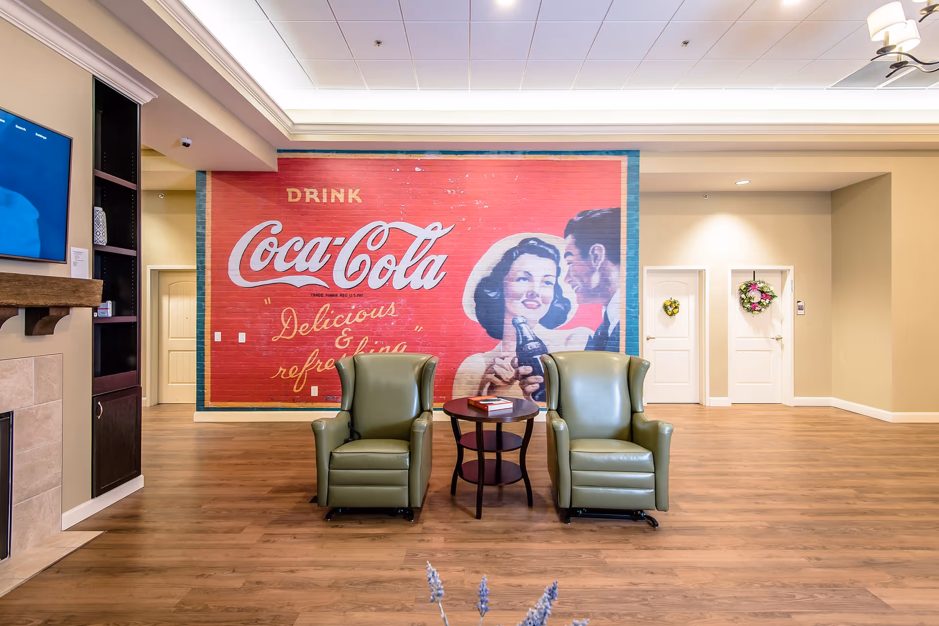 A cozy seating area in a senior living facility with two green armchairs and a small round table between them. Behind the chairs is a large vintage Coca-Cola mural on the wall featuring a man and woman holding a bottle of Coca-Cola. The room has wooden flooring, beige walls, and two white doors decorated with floral wreaths. A flat-screen TV is mounted on the left wall above a fireplace.