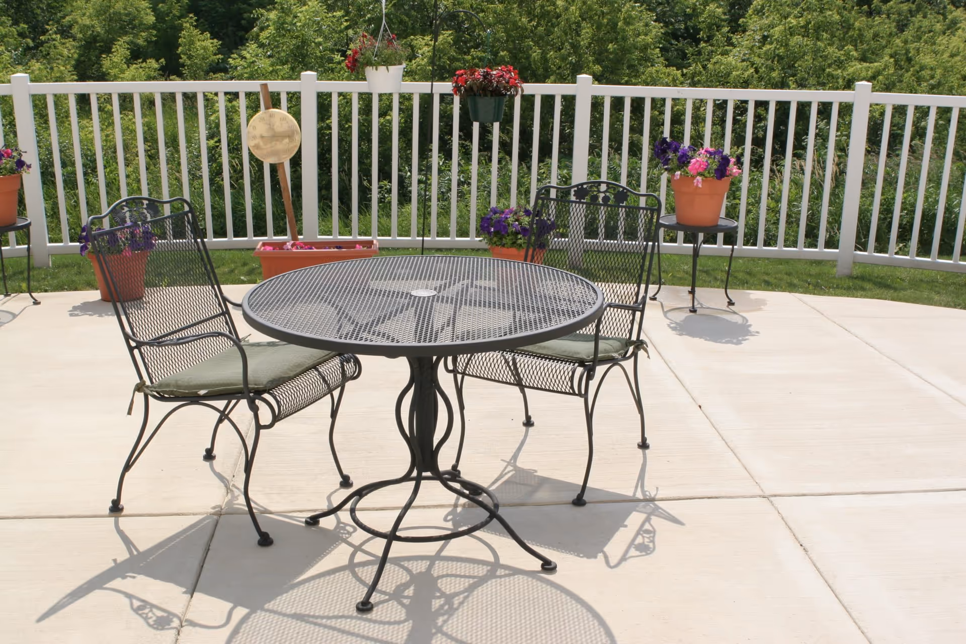Outdoor patio area with a round metal table and two metal chairs with green cushions. The patio is surrounded by a white fence and decorated with several potted and hanging flowers. Green trees and grass are visible beyond the fence.